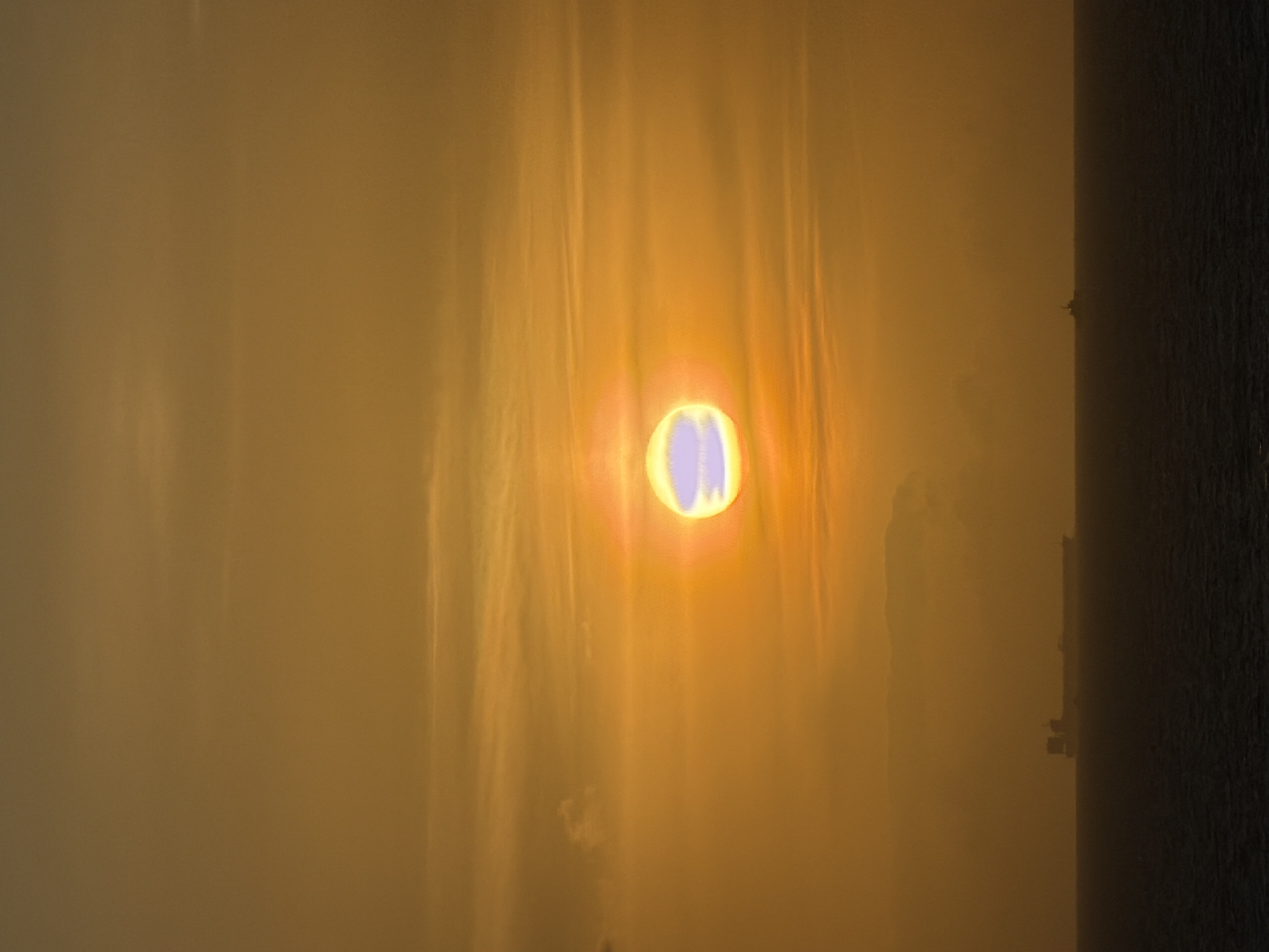 A hazy view shows a sun-like light source with a bluish center, casting vertical beams of light through what appears to be a polluted or foggy environment. A dark, textured structure occupies the right edge of the image. The location seems indeterminate.