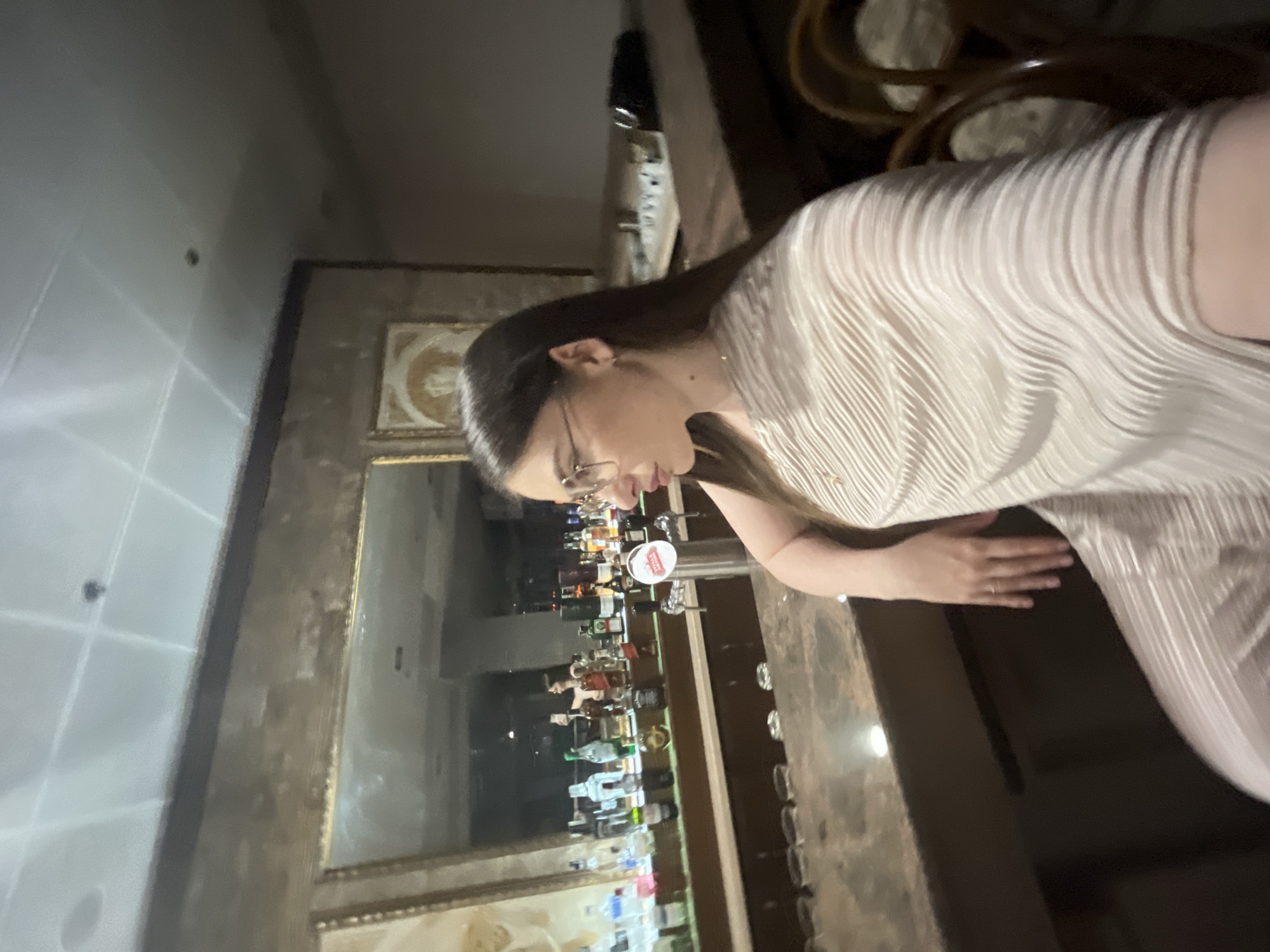 A woman wearing glasses and a light-colored, textured dress is standing at a bar with a mirror reflecting a display of liquor bottles. The location appears to be in a city, but the specific name and location of the city are unknown. The lighting is dim.