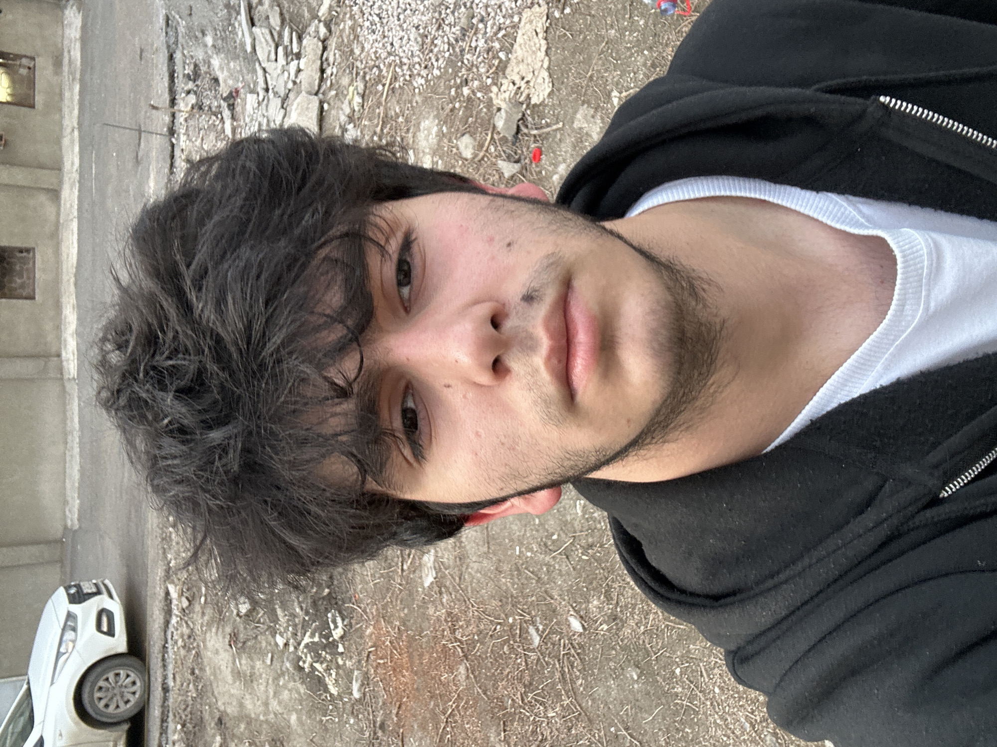 A young man stands outside, facing the camera directly. He has dark hair, and stubble on his face. He is wearing a white t-shirt under a black zip-up hoodie. The ground appears to be a mix of dirt, gravel, and debris. In the background, there is a gray building and a white car. The location is a city street.