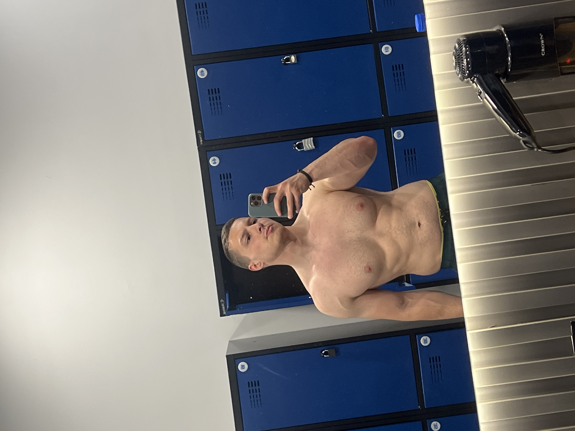 A shirtless Caucasian male is taking a selfie in a locker room. The background includes blue lockers and a hair dryer. The man is wearing a bracelet. The location is Burgas, Bulgaria.