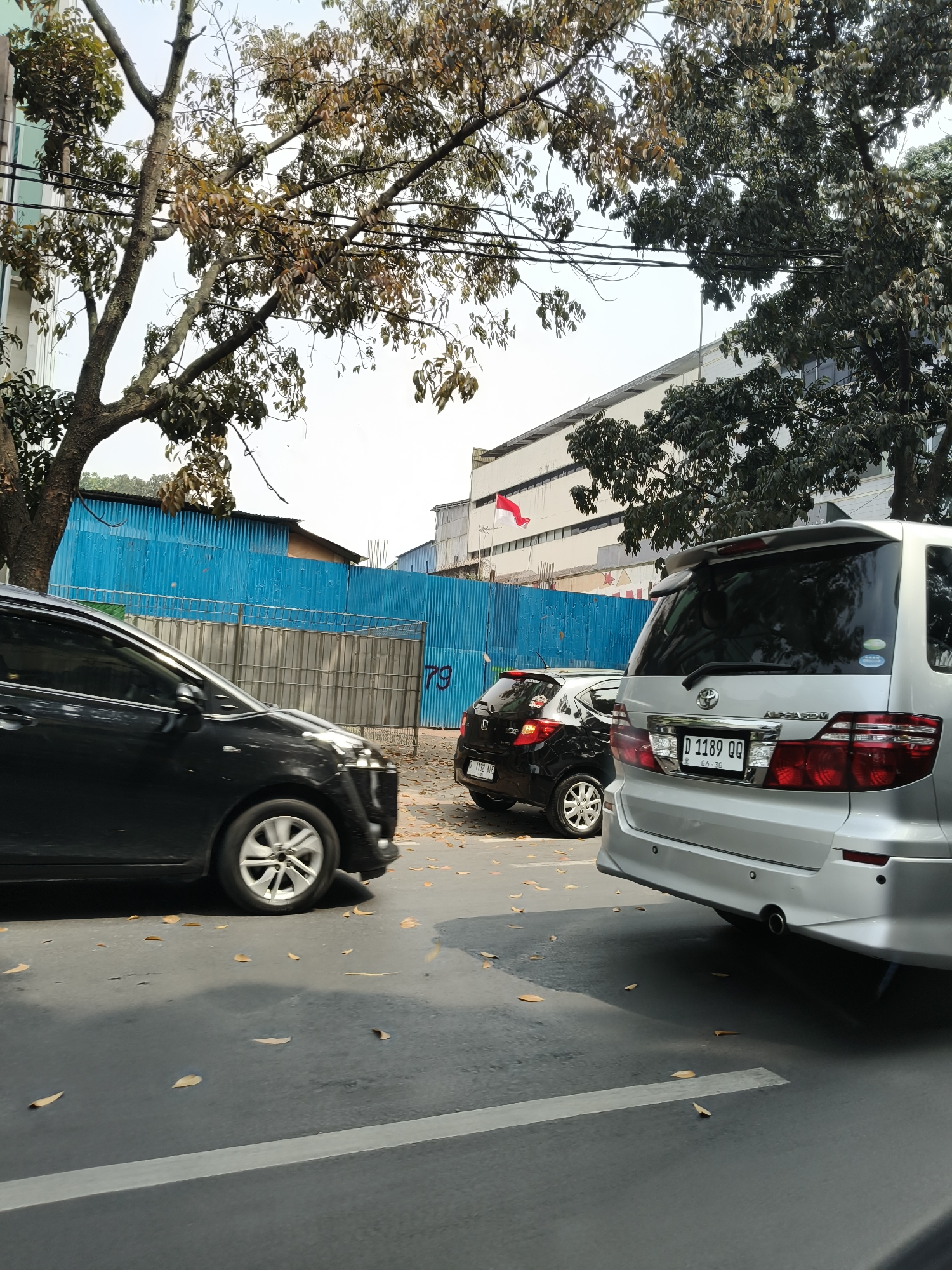 The image depicts a street scene with several cars parked or moving. In the foreground, a silver minivan is parked on the right, with its rear facing the camera. To its left, a black sedan is partially visible, also from the side. Further down the street, a small black hatchback is parked. Behind the cars, a blue corrugated metal fence runs horizontally, likely a construction barrier. A building with white and grey panels is visible behind the fence, and a small Indonesian flag is flying on a pole in front of it. The street is paved, with fallen leaves scattered on the asphalt. The overall setting appears to be an urban environment, possibly in Bandung City, Indonesia, given the Indonesian flag. The lighting suggests it is daytime, with sunlight casting shadows on the road.