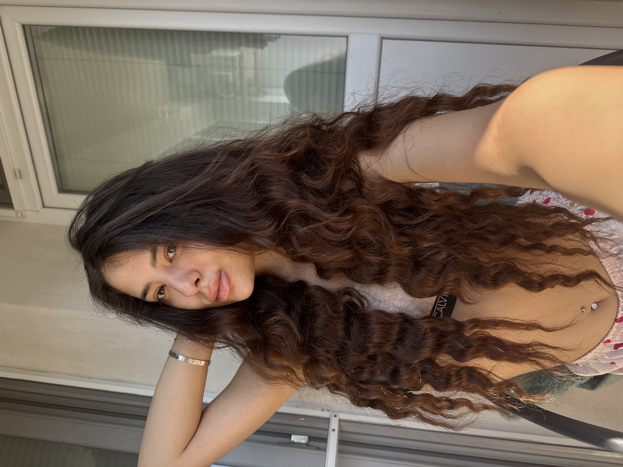 A young woman with long, dark, curly hair stands outdoors, leaning against a balcony railing. She is wearing a light-colored bra with a Calvin Klein logo band and pajama bottoms. She has a bracelet on her left wrist and a belly button piercing. Sunlight illuminates her face and arm. In the background, a white sliding glass door with blinds is visible. The scene is likely a residential balcony in Plovdiv, Bulgaria, given the architectural style of the building and the presence of a balcony.