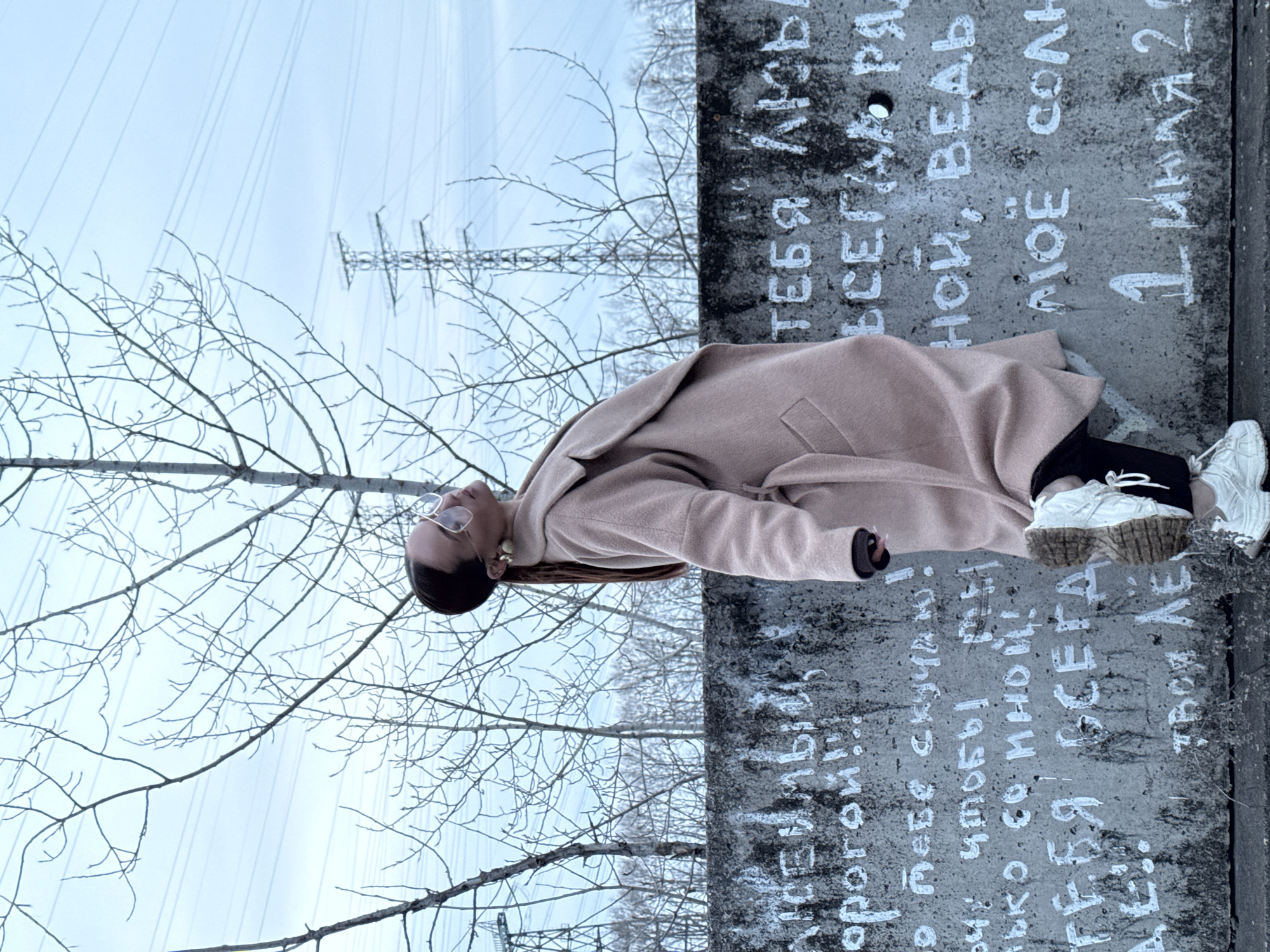 A young woman wearing a beige coat and white sneakers is reclining on a concrete structure with graffiti written in Russian. The graffiti includes phrases like "Love you always" and "Always with you." The date "July 1, 202_" is also visible. In the background, bare tree branches reach towards the sky, and an electrical transmission tower stands in the distance. The scene appears to be an outdoor setting in the city of Chaikovsky, Russia.