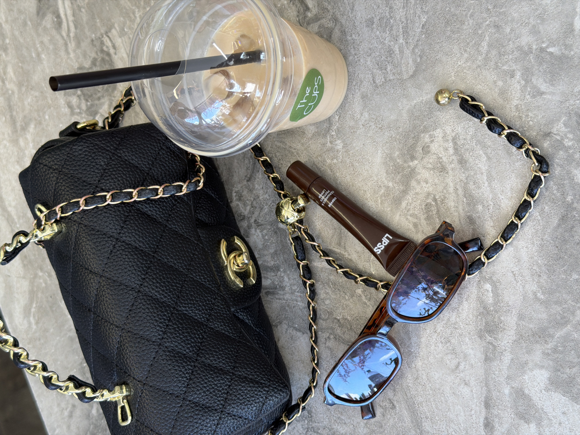The image displays a flat lay arrangement of fashion accessories and a beverage on a speckled gray surface, possibly a stone countertop. In the upper left quadrant, a black quilted handbag with a gold chain strap is prominent. To the right of the handbag is a clear plastic cup filled with an iced coffee beverage, a black straw, and a green sticker on the cup that reads "The CUPS". In the lower half of the image, a pair of tortoiseshell sunglasses rests near a small, dark brown tube with the label "LIPSS" visible. The gold chain from the handbag is draped across the sunglasses and the lip gloss tube, with a small gold charm attached to the chain. The lighting suggests an indoor setting, possibly a cafe or outdoor seating area. No specific landmarks or architectural features that would pinpoint a location within Kamianske, Ukraine are visible.