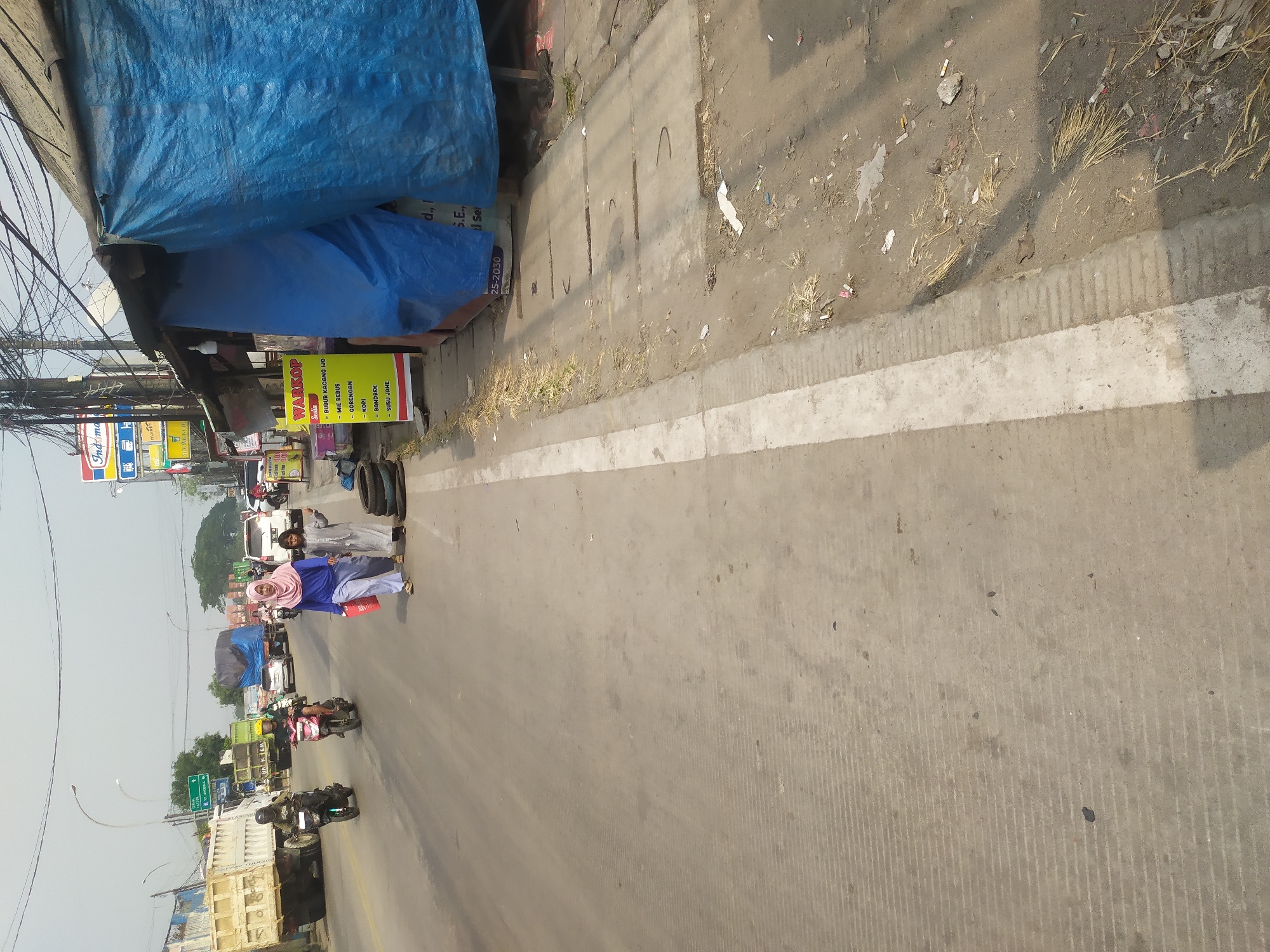 In Kragilan, Indonesia, on a bright, hazy day, a street scene unfolds. A two-lane road, marked with a white stripe down the center, is busy with traffic. On the left lane, two motorcycles are moving away from the viewer, their riders visible. Further down the road, a large, white dump truck is parked. On the right side of the road, a woman in a pink hijab and a light blue outfit walks towards the viewer, carrying a red bag. Beside her, a child in a grey outfit walks in the same direction. Behind them, several stacked tires are visible near a small, open-air establishment with a blue tarp covering its roof and side. A yellow sign with red lettering, possibly a menu or advertisement, stands in front of the establishment. The ground is a mixture of asphalt and dirt, with scattered debris and dry grass. Power lines crisscross overhead. The overall impression is one of a bustling, everyday scene in a developing urban area.