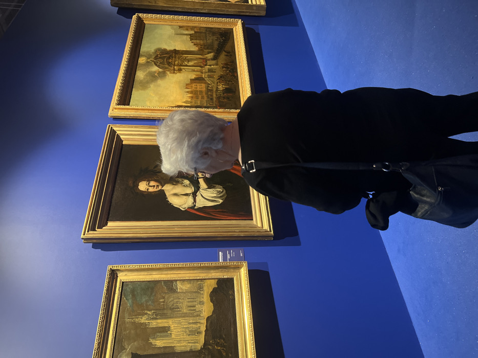 An art gallery or museum in Unknown, Unknown. An elderly individual, observed from behind, with short white hair and glasses, wearing a black long-sleeved top and black trousers, stands facing a vibrant blue wall. A black shoulder bag with a silver buckle strap hangs on their left side. Three framed oil paintings are displayed on the blue wall. From left to right: the first painting is a vertical architectural landscape depicting a large, light-colored Gothic-style building under a dark sky. The central painting is a vertical portrait of a woman with dark hair, a pale complexion, wearing a white off-the-shoulder garment and a red cloak, with visible chains on her wrists. The third painting is a vertical cityscape showing a large crowd in a square, with a prominent domed structure emitting smoke. The floor is covered in a matching blue material. Small informational plaques are visible below the leftmost and central paintings.