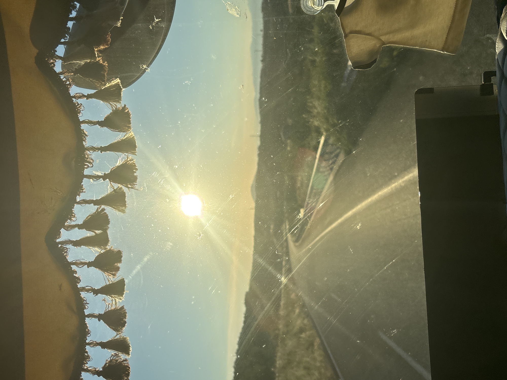 The image shows a view from inside a vehicle, looking out through the windshield. The sun is setting, casting a warm glow over a winding road that leads into a hilly landscape. The sky is a clear blue, with the sun positioned in the upper center of the frame. A decorative fringe with tassels hangs from the top of the windshield. In the foreground, a dark dashboard is visible, and a light-colored t-shirt is hanging from a hook on the right side of the windshield. The location is identified as Avra, Greece.