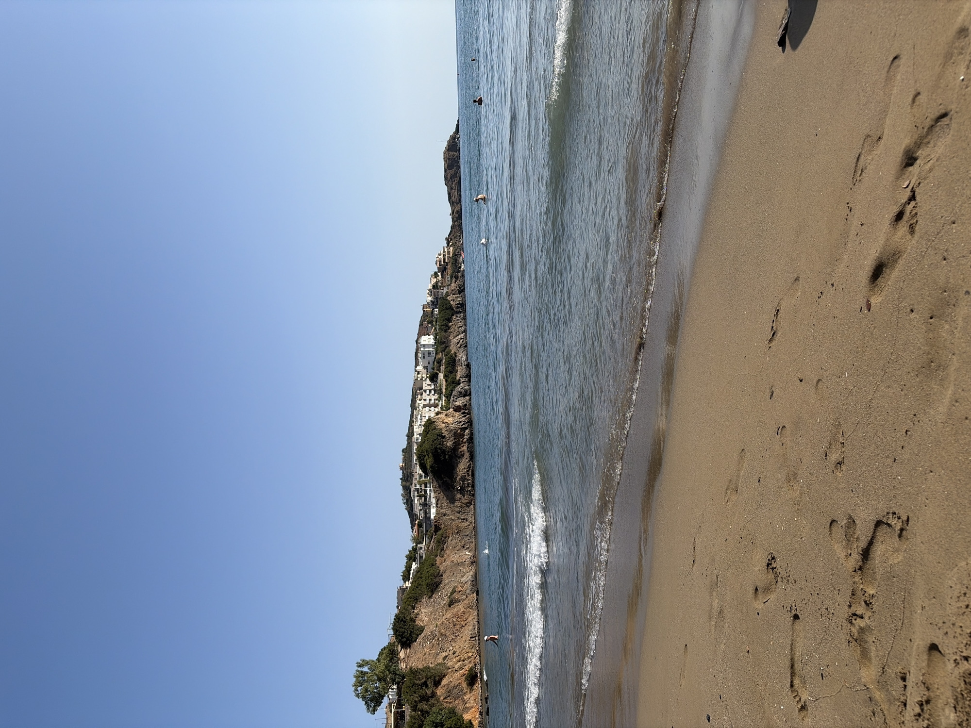 The image depicts a sunny day at a beach in Vlichada, Greece. The foreground is dominated by a sandy beach with visible footprints, indicating recent human activity. Gentle waves lap the shore, creating a white foam line along the wet sand. In the middle ground, the calm, blue sea stretches towards the horizon. Several individuals are visible in the water, some swimming and others standing. In the background, a rocky coastline rises, dotted with green vegetation and a cluster of white buildings, characteristic of coastal architecture in Greece. The clear blue sky above completes the serene beach scene.