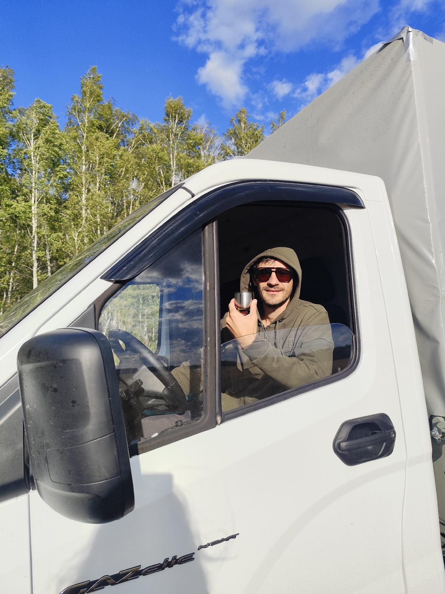 A man wearing sunglasses and a hooded jacket is sitting in the driver's seat of a white truck. He is holding a small metallic cup in his right hand and looking at the camera. The truck is parked in an outdoor setting with trees and a blue sky with white clouds in the background. The scene appears to be in an rural area, possibly on a road or in a parking lot. The logo on the truck indicates it is a Gazelle. The surrounding area is a rural landscape with trees and a clear blue sky. The overall impression is a snapshot of a person taking a break during a journey.