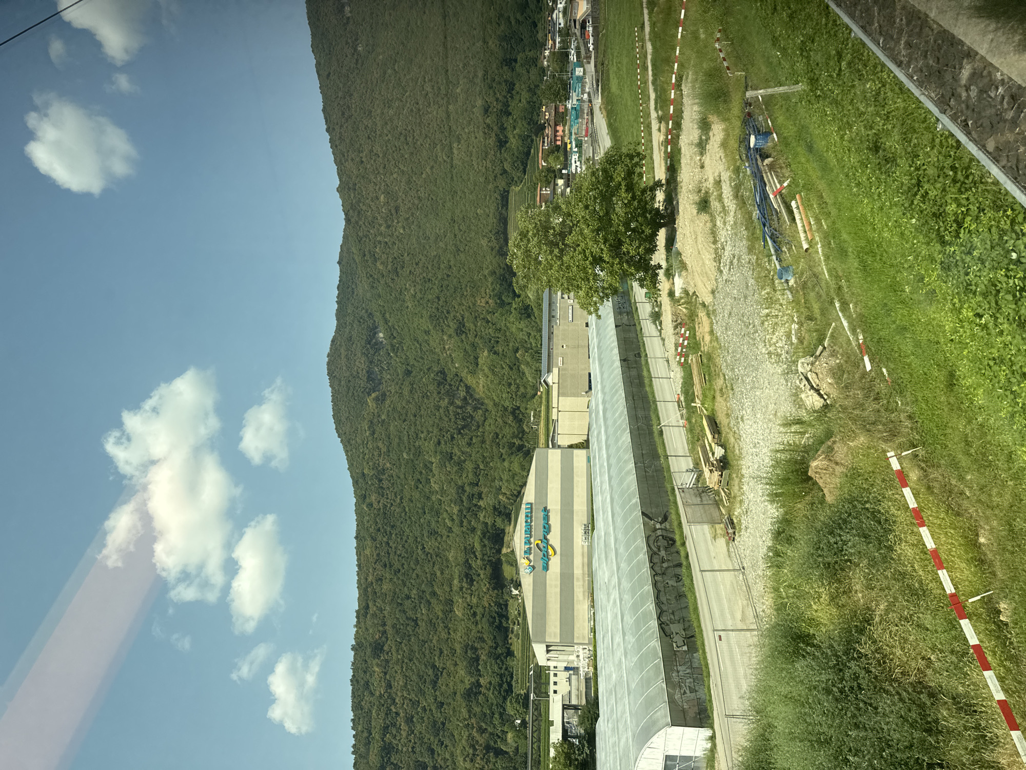 In Riva San Vitale, Switzerland, a wide shot captures an industrial area nestled against a verdant, tree-covered hillside under a partly cloudy blue sky. In the foreground, a grassy slope is marked by red and white striped caution tape. Further back, a large, long, white-roofed structure, possibly a greenhouse, is visible, its side covered in graffiti. Adjacent to it stands a two-story building with a gray and beige facade, bearing the logos "V. PURICELLI" and "alpriget" in blue and yellow. A mature, leafy green tree stands prominently in the middle ground. Beyond this, a small town with red-tiled roofs is scattered along the base of the hill, with a road and more industrial buildings visible. Various construction materials, including pipes and lumber, are present in the open, undeveloped areas.