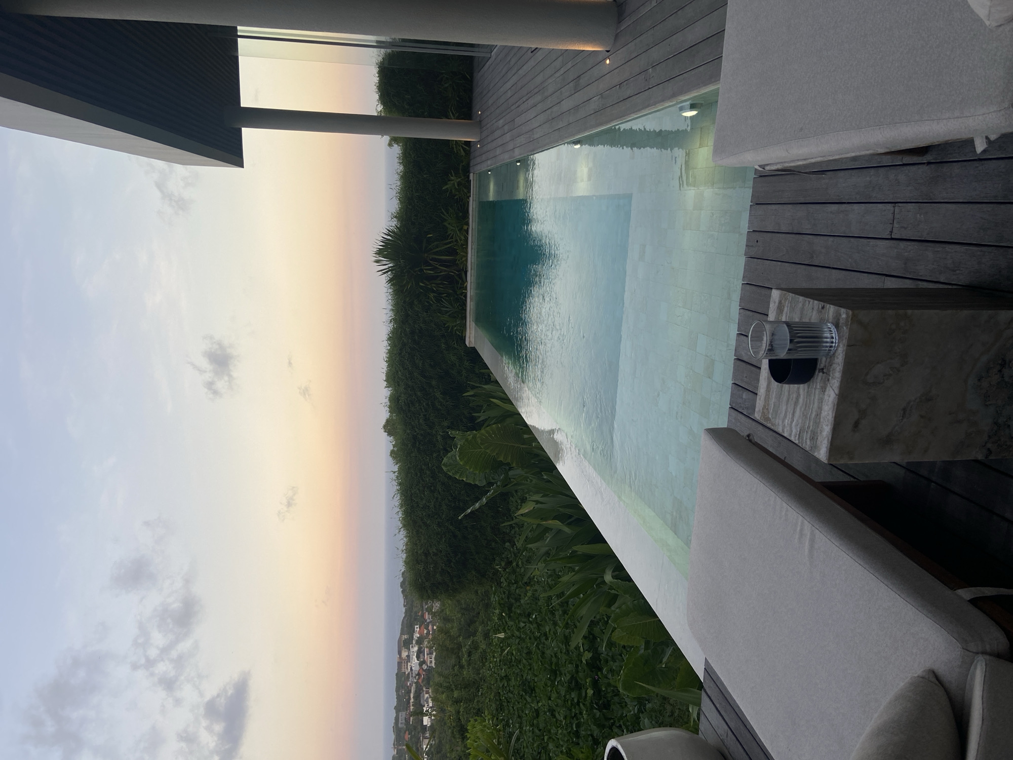 The image shows a view from a luxurious outdoor area with an infinity pool. The location appears to be in a hilly area, possibly near the city of Kuta Selatan, Indonesia, based on the vegetation and the distant view of what seems to be a coastal cityscape. The sky is a gradient of colors, suggesting either a sunset or sunrise. A clear glass containing a dark-colored liquid is on a table next to the pool. There are lounge chairs near the pool, offering a space for relaxation. The area is surrounded by dense greenery and some buildings, blending with the natural environment.