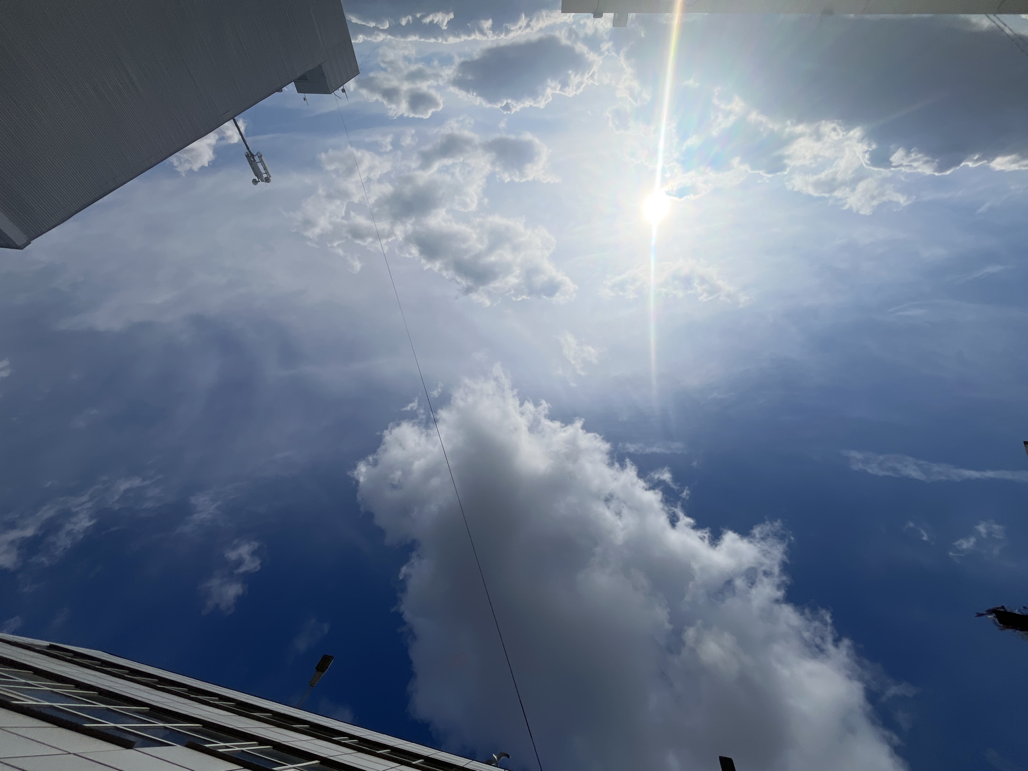 The image shows a low-angle view of the sky in Klin, Russia. Predominantly blue sky is partially obscured by white clouds. The sun is high in the sky. Building edges appear at the top-left and bottom-left of the frame. A suspended maintenance platform hangs from a cable descending from the top-left building edge. A street light is visible near the building in the bottom-left corner.
