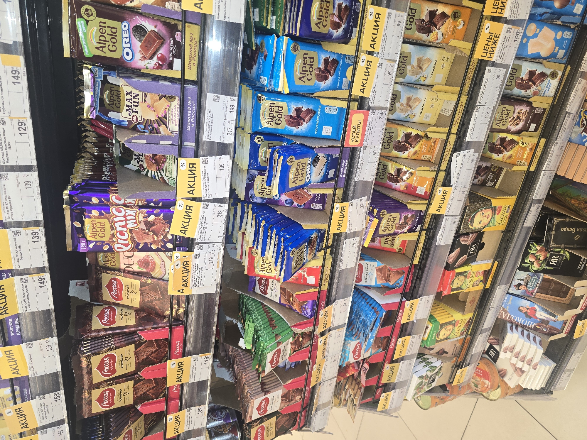The image shows a brightly lit supermarket aisle stocked with various chocolate bars and confectionery products. The shelves are organized, displaying a wide selection of brands and flavors, many of which are marked with promotional "АКЦИЯ" (sale) tags in yellow. Prominent brands include "Россия" and "Alpen Gold," with packaging featuring images of chocolate, fruits, and nuts. Some products are displayed in cardboard risers. The overall environment suggests a retail setting, likely a grocery store or supermarket, with clean aisles and well-maintained shelving. There are no people visible in the frame. The specific location in Ulan-Ude, Russia, cannot be determined solely from the visual information in the image.