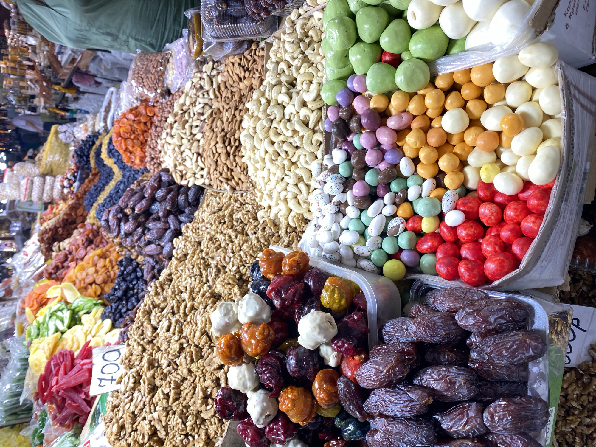 The image depicts a vibrant market stall in Makhachkala, Russia, overflowing with a colorful assortment of dried fruits, nuts, and candies. The stall is densely packed, with various items arranged in piles and containers. In the foreground, a large quantity of dates, a pile of walnuts, and an assortment of brightly colored candies in shades of red, white, orange, purple, green, and speckled hues are prominently displayed. Behind these, a substantial heap of almonds and cashews is visible, along with dried apricots and other dried fruits in the background. The overall impression is one of abundance and variety, typical of a bustling marketplace.