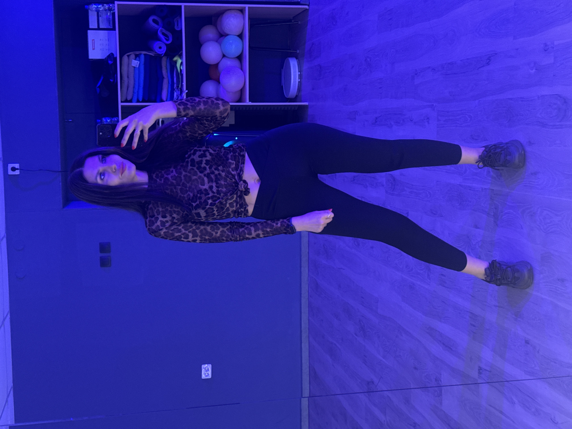 The image shows a young woman standing in what appears to be a gym or dance studio, reflected in a large mirror. She is wearing a leopard-print long-sleeved mesh top tied at the waist and black leggings. She is holding a smartphone in her right hand, taking a selfie. The room is dimly lit with blue ambient lighting, which illuminates a wooden floor and a wall with electrical outlets and light switches. In the background, there is a shelving unit with various items, including folded towels, yoga mats, and several spherical objects, possibly exercise balls. The woman has long dark hair and is wearing dark sneakers. There is no indication of the city of Chaikovsky, Russia, in the image.