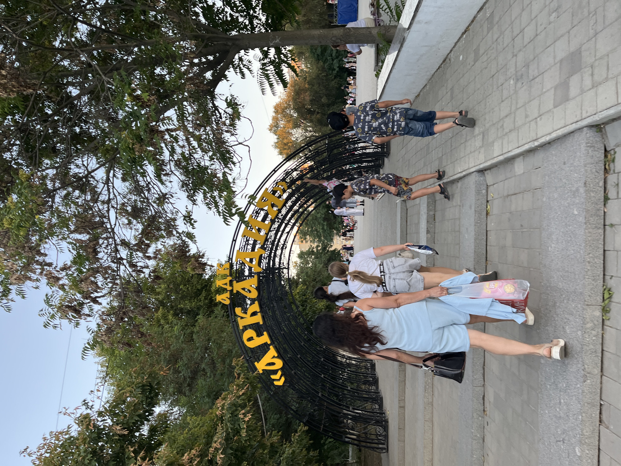 The image depicts an entrance archway to the "Arkadiya" park in Astrakhan, Russia, an amusement park. The arch is made of black metal with yellow Cyrillic lettering spelling out "Arkadiya." Several people are walking through and towards the arch. In the foreground, a woman in a light blue romper is walking up a set of stairs with a black purse and a plastic bag containing clothing and snacks. Behind her, another woman in denim shorts and a white t-shirt walks up the stairs. Two young boys in patterned t-shirts and shorts are also visible walking up the paved pathway. Trees with green and dry leaves surround the park entrance, and the sky is clear and blue. In the background, more people are seen inside the park, along with buildings and trees. The scene suggests a warm, late afternoon or early evening in a public recreational area.