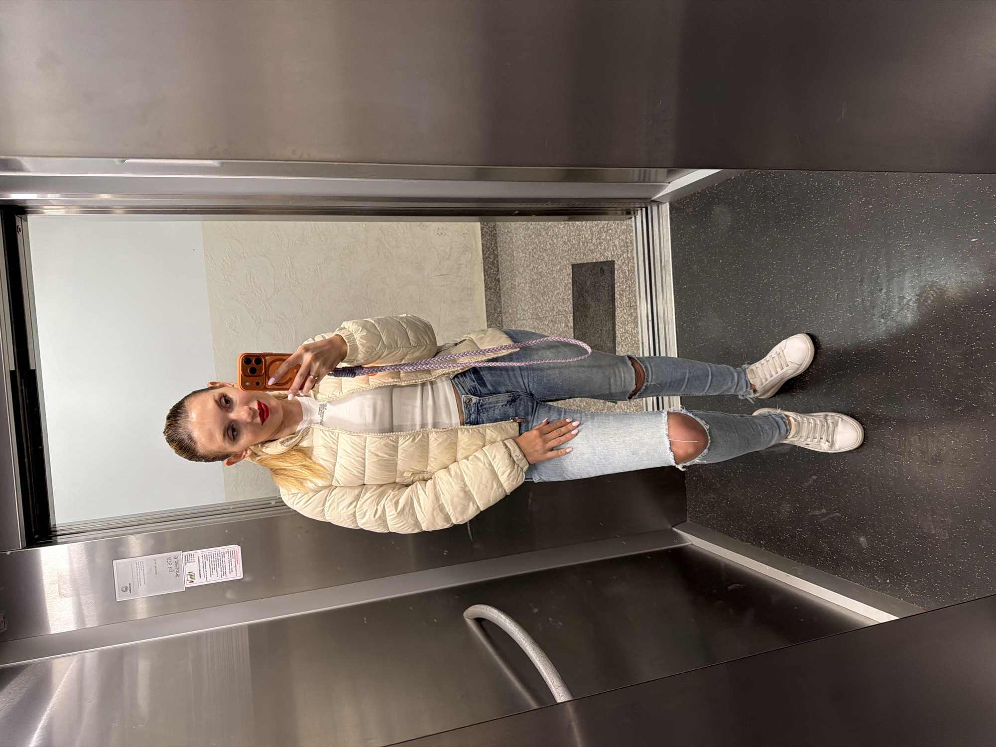 A woman takes a selfie in an elevator. The elevator has stainless steel walls and a black speckled floor. The woman is wearing a white puffer jacket, a white top, ripped blue jeans, and white sneakers. She is holding an orange smartphone. The reflection outside the elevator shows a beige wall and a gray tile floor. There is no information available about whether this image was taken in Budva, Montenegro.