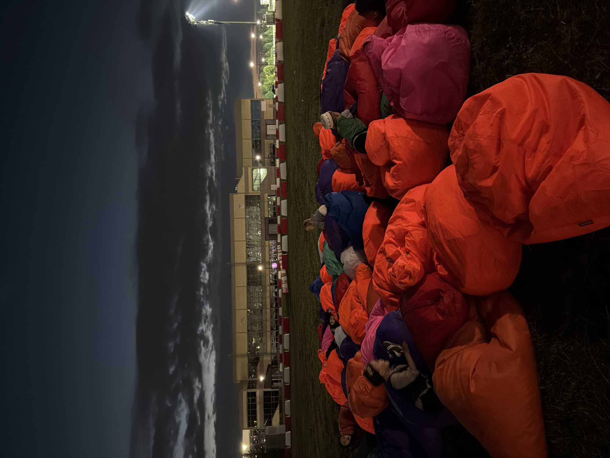 A group of people are sitting on a large pile of colorful bean bag chairs on a grassy field at dusk. In the background, there is a building with many windows and a bright stadium light. The sky is dark with some clouds. Some people are looking at the camera, while others are looking in different directions.