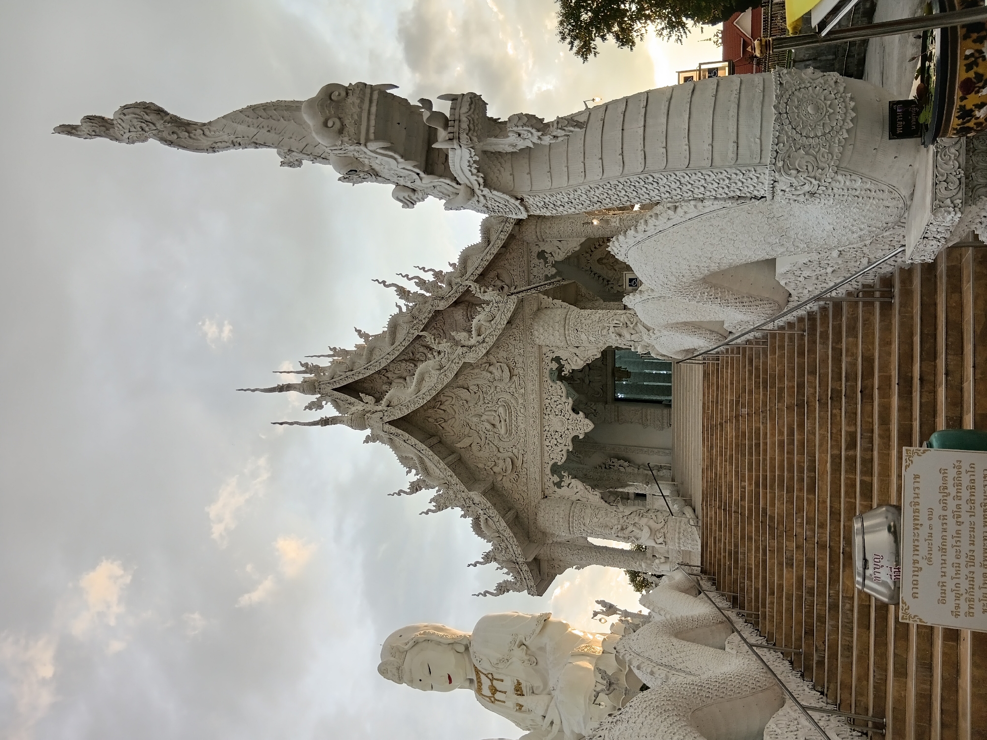 The image shows a grand white temple in Baan Lao Phatthana, Thailand. The temple features intricate architectural details, a steep staircase leading to its entrance, and large white dragon statues flanking the stairs. To the left of the stairs, a large white statue of a seated figure, possibly a deity, is visible. The sky above is cloudy with hints of sunset light. In the foreground, there is a sign with Thai text and a metallic receptacle at the base of the stairs. The overall scene conveys a sense of spiritual and architectural grandeur.