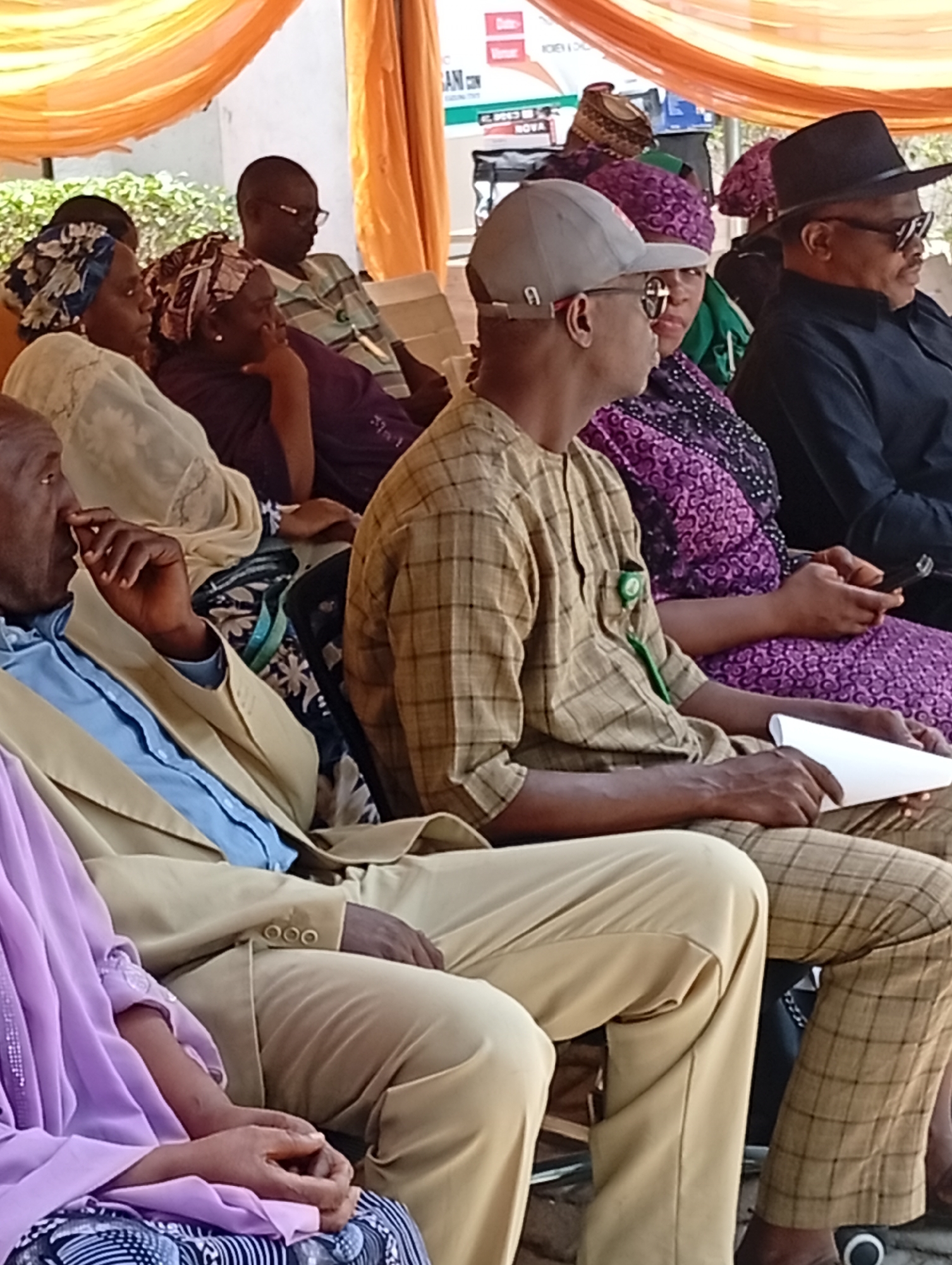This image depicts a daytime gathering of people, primarily adults, in Unguwar Sarki, Nigeria, likely an outdoor or semi-outdoor event. Bright natural light illuminates the scene, with decorative orange fabric draped overhead, suggesting a temporary setup.
The subjects are seated in chairs, appearing attentive. Their attire is a blend of traditional Nigerian garments and more casual wear. Notable individuals include:
* A man on the far left, partially visible, wearing a light blue collared shirt beneath a tan suit jacket, with his hand near his face.
* Next to him, a person is seen in a vibrant light purple traditional outfit.
* In the foreground, a man wears a beige and brown plaid shirt, a grey baseball cap worn backwards, and sunglasses. A green lapel pin is affixed to his shirt, and he holds a rolled white paper.
* Beside him sits a woman dressed in a purple patterned outfit with a matching head covering, holding a mobile phone.
* Further right, a man with a dark complexion wears a black fedora-style hat, sunglasses, and a black shirt.
* Other attendees are visible in the background, including women wearing patterned head coverings and a man in glasses with a striped shirt.
The activity taking place appears to be a formal or semi-formal meeting or event, given the seated arrangement and the focused demeanor of the participants. There are no strong emotions visible, with most individuals appearing calm and engaged. In the background, a banner is partially visible with text that includes "ICAN CON" and "NOVA", possibly indicating the name of an organization or the event. The overall scene suggests a community gathering or a public event.