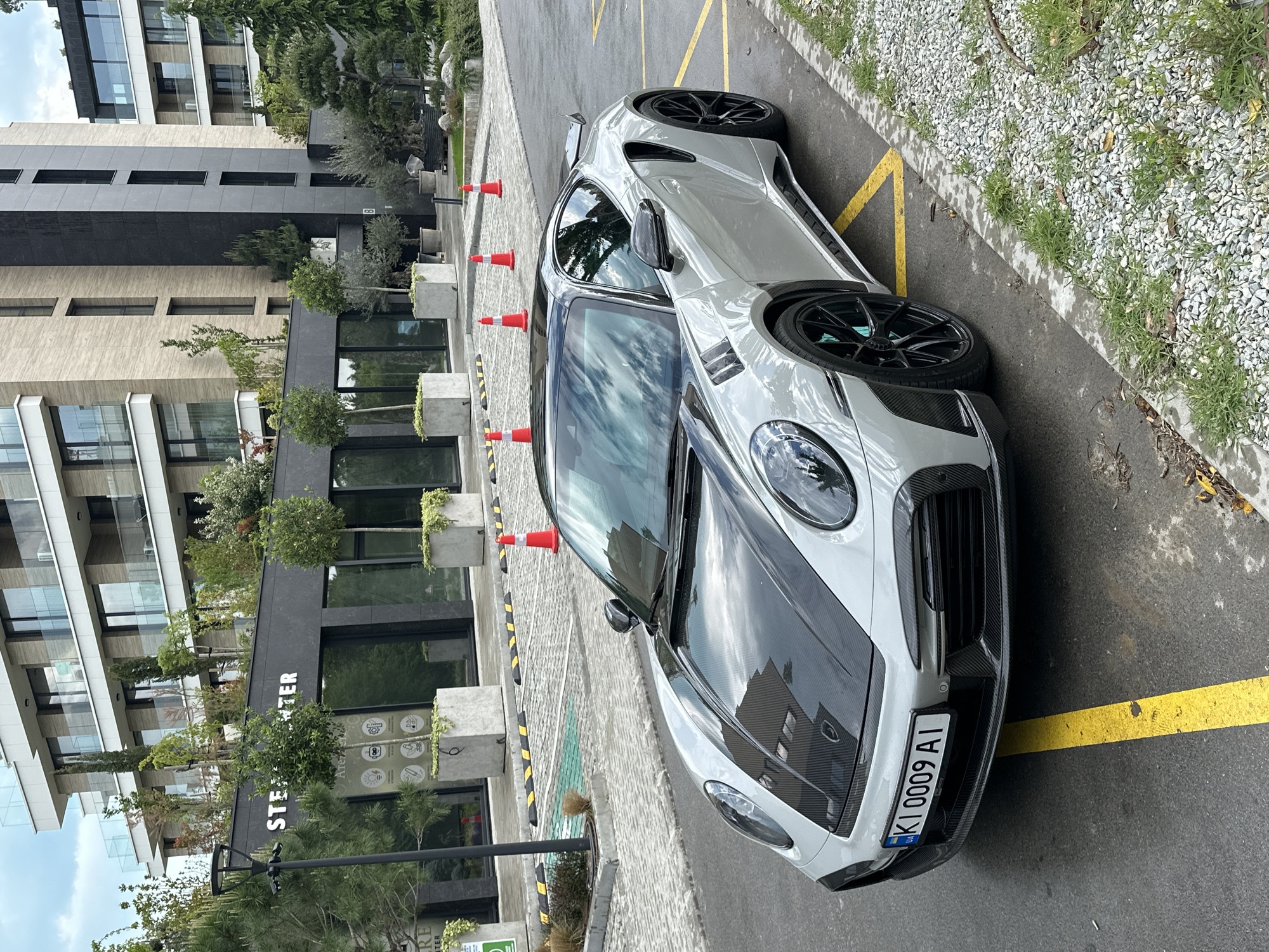 The image depicts a silver Porsche parked on a paved surface. Orange cones are placed nearby. The background includes a modern building with "STEARTER" signage and greenery. The license plate on the car reads "KI 0009 AI". The location is likely urban, possibly a parking area in Varna, Bulgaria.