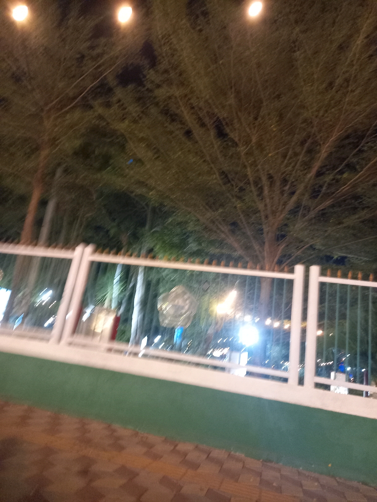 The image depicts an outdoor scene at night, likely in Pamekasan, Indonesia, characterized by trees with dense foliage and illuminated by artificial lights. A white fence with vertical green bars and some yellow conical spikes adorns the foreground. Beyond the fence, blurred lights and foliage suggest a park or public space. The ground in the immediate foreground is paved with reddish-brown rectangular stones arranged in a herringbone pattern. The overall impression is one of a dimly lit, possibly recreational area at night.