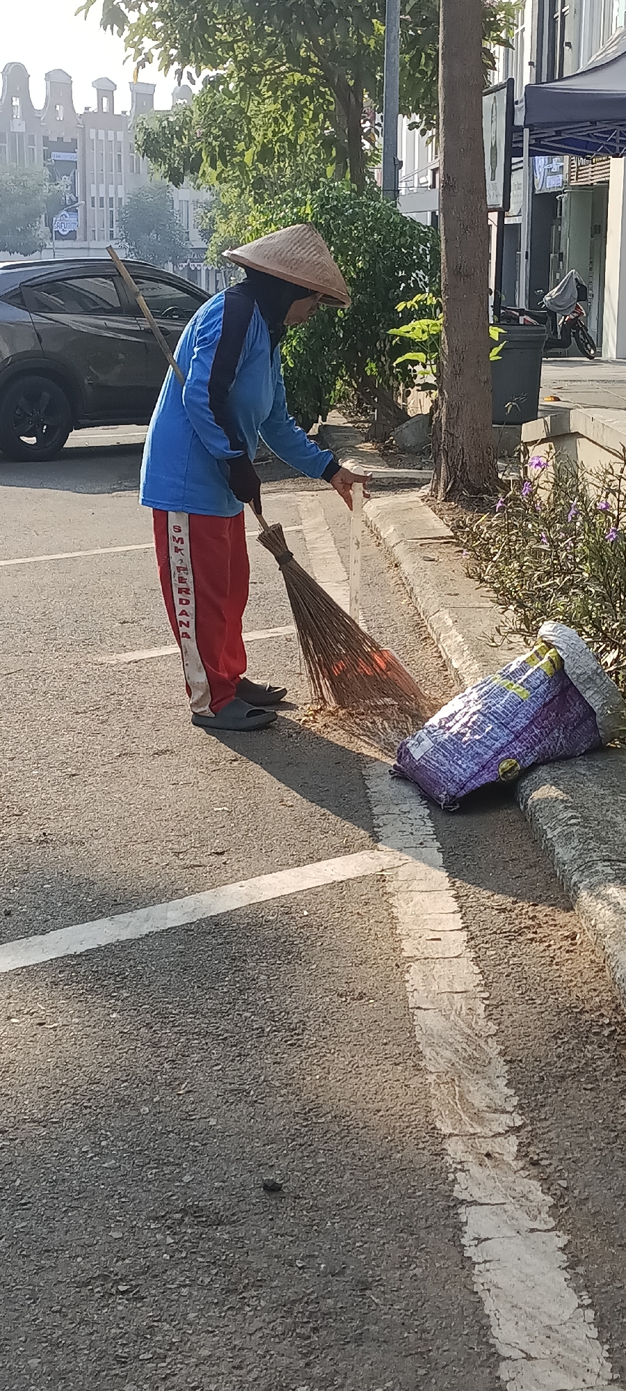 An individual, wearing a blue long-sleeved shirt, red pants with white stripes, a black headscarf, and a conical straw hat, sweeps debris from a paved area with a broom. A bamboo stick is propped on their shoulder. To the right, a large, partially visible purple and silver plastic bag lies on its side, and beyond that, a patch of purple flowers and a tree trunk are visible. A grey car is parked to the left, and buildings can be seen in the background. The scene takes place on a street in Semarang, Indonesia, indicated by the architectural style of the background buildings which resemble those found in Kota Lama Semarang, the old town.