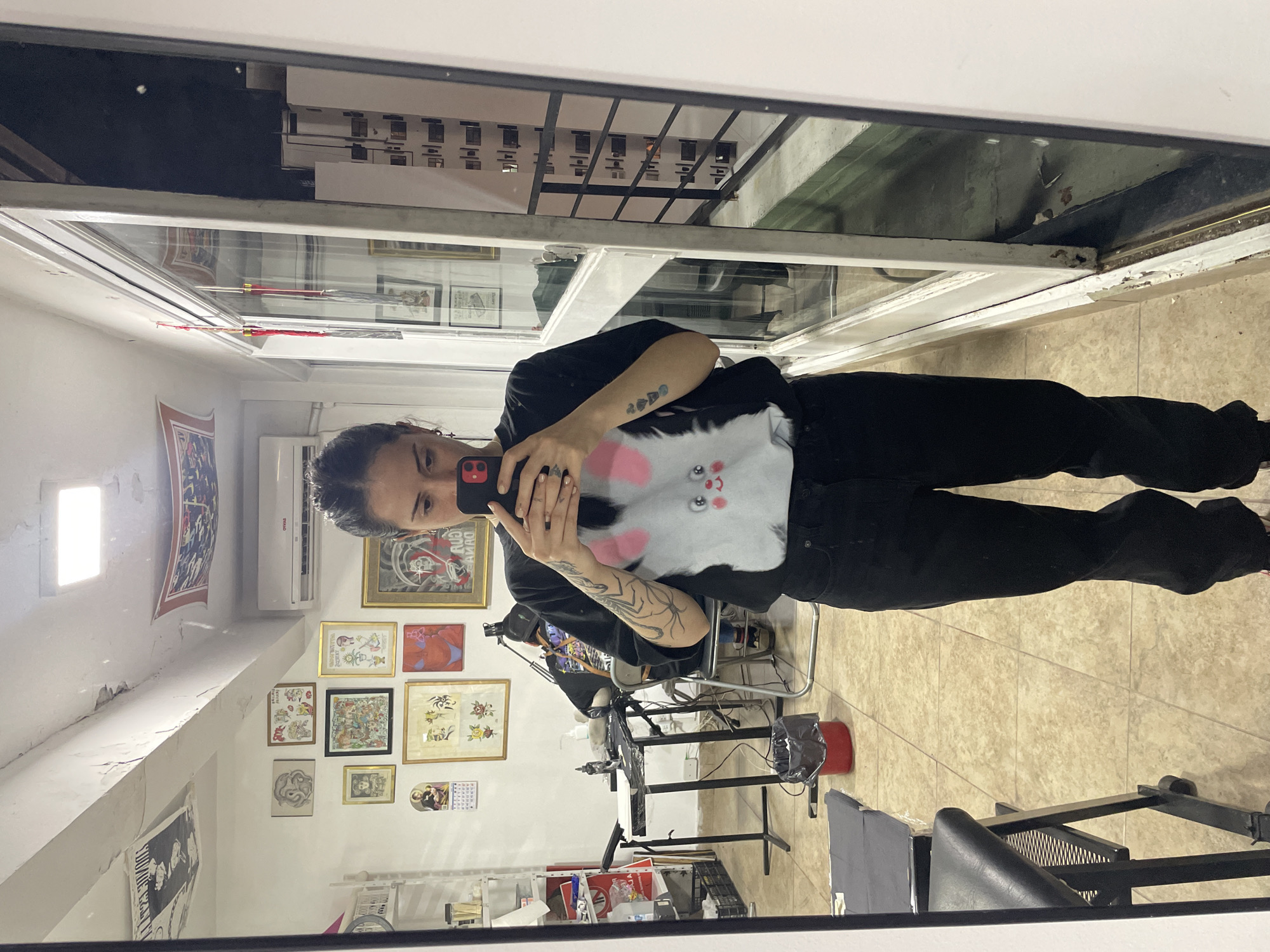 A woman is taking a selfie in front of a mirror. She's wearing a black t-shirt with a graphic of a bunny face and black jeans. The background appears to be a tattoo studio, with framed tattoo designs on the wall. There's a tattoo workstation with equipment visible. Outside the window, buildings are visible. The location may be the city of Buenos Aires, Argentina.