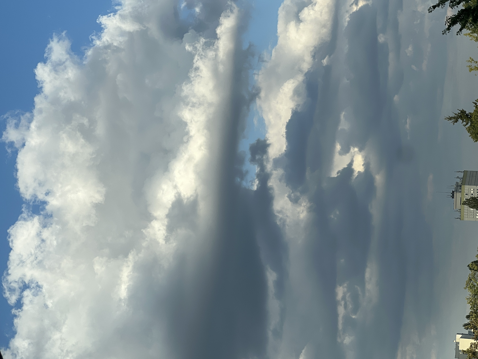 The image captures a view of the sky above Warsaw, Poland, with dramatic, layered clouds. The sky is a bright blue with scattered sunlight breaking through the dense cloud formations. The clouds range in color from bright white to dark gray, indicating varying thickness and density. Towards the bottom of the image, the upper portions of several buildings and trees are visible, suggesting an urban or suburban environment. The buildings appear to be modern in design, with one prominent structure featuring multiple antennas on its rooftop. The overall scene depicts a dynamic and atmospheric sky over a cityscape.