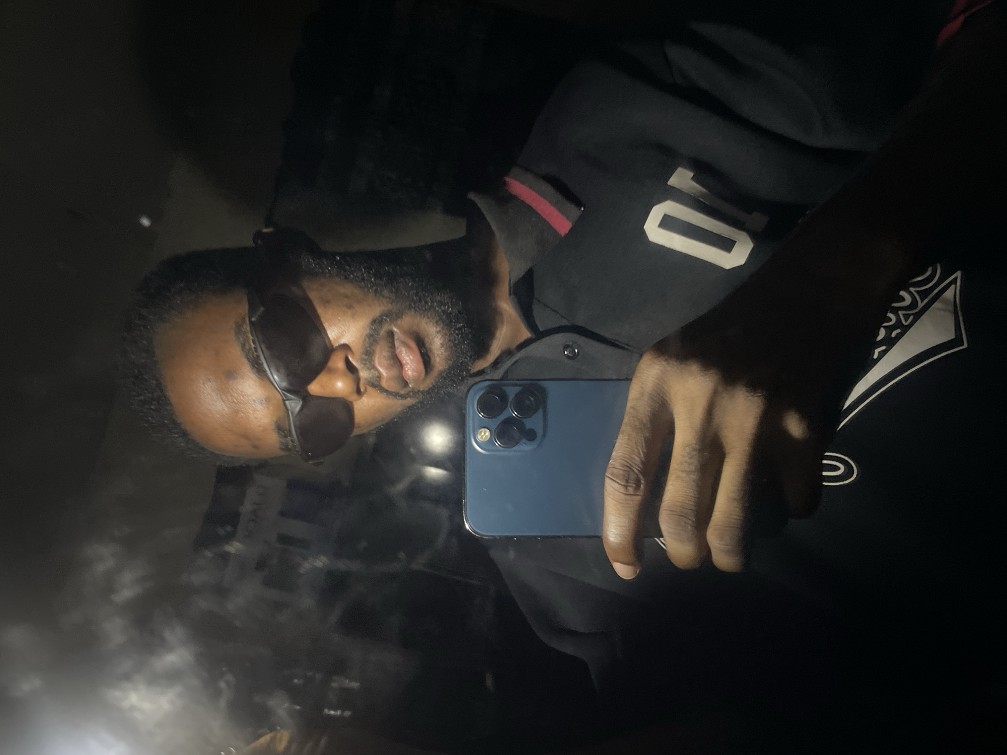 An adult male with dark skin and a short beard is depicted taking a selfie. He wears dark sunglasses and a black polo shirt with a black collar featuring a red stripe. A white numeral, possibly "0" or "O", is present on the chest of his shirt, and a white patterned design is visible on the lower left portion of his garment. He holds a blue smartphone, oriented vertically, in his right hand, with the device's multi-lens rear camera visible. The image is captured in a dark, reflective environment, likely a vehicle interior or a room with a mirror or window at night. The background features blurred reflections of an urban setting, including discernible building structures and ambient light sources. Faint, indistinct text and shapes, characteristic of building signage, are reflected in the upper left portion of the background. The scene is located in the city of Lokoja, Nigeria.