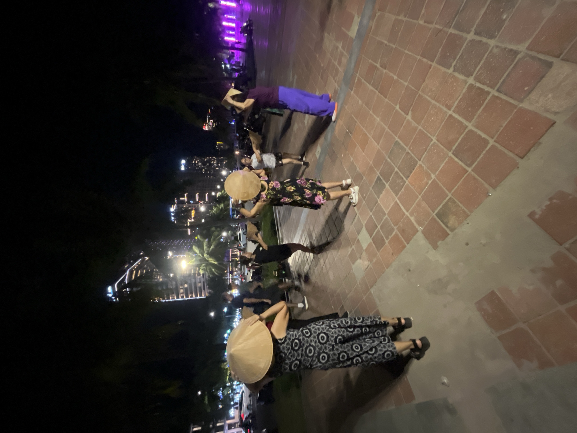This image shows a group of people on a brick-paved walkway at night in Da Nang, Vietnam. A tall building with many illuminated windows is visible in the background, along with palm trees and streetlights. Several individuals are wearing conical hats and appear to be engaged in a dance or performance, holding their hats as props. One woman in the foreground is wearing a black and white patterned dress and sandals, while another person in the middle ground wears a floral dress. To the right, a person in a purple top and blue pants also holds a hat. The scene is illuminated by artificial lights, creating a vibrant nighttime atmosphere in an urban setting.