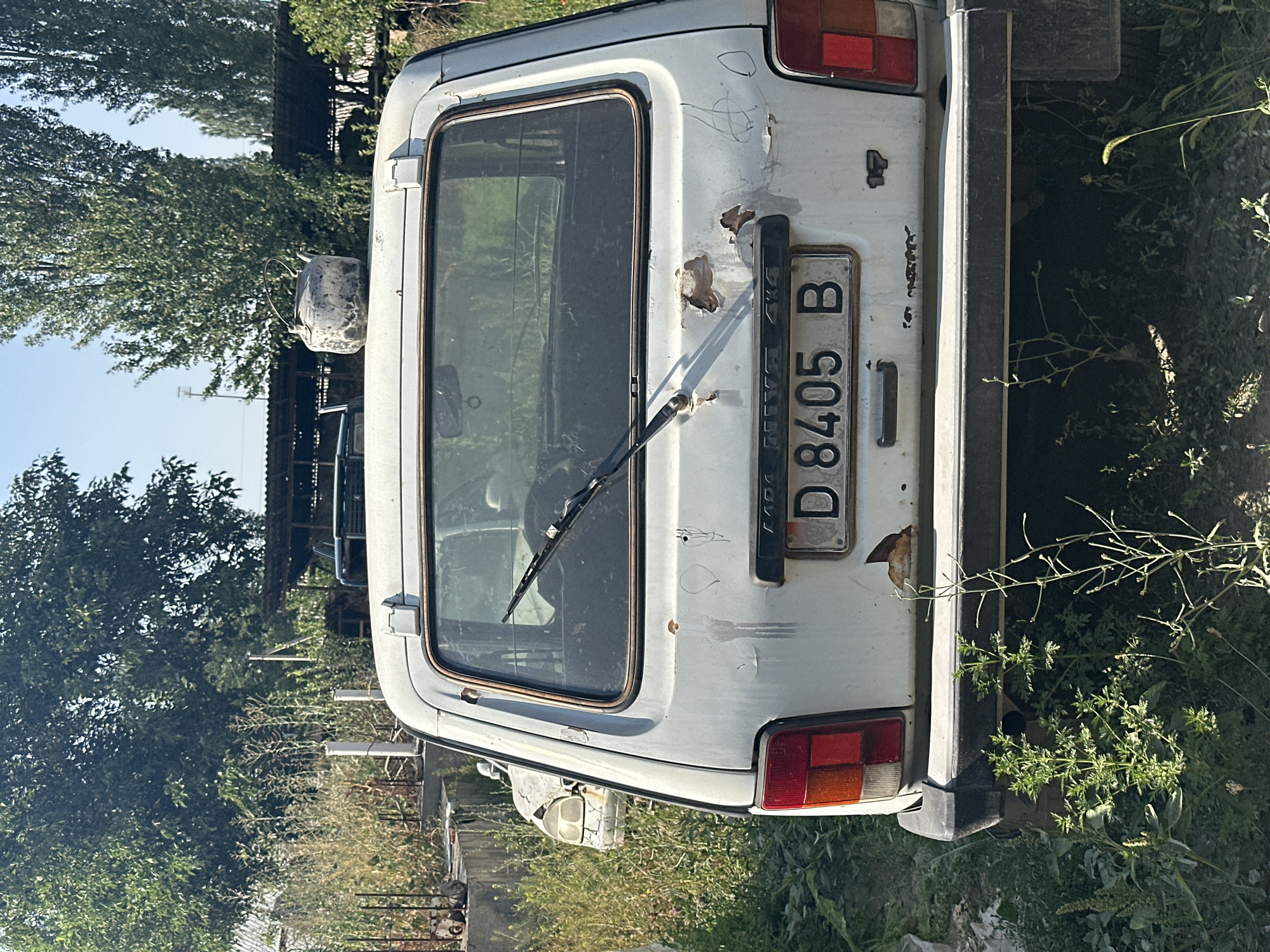 A white, older model Lada Niva 4x4 is parked outdoors in an overgrown lot, likely in International, Kyrgyzstan. The car is rear-facing, with a license plate reading "D 8405 B". It appears weathered and rusty, with some of the paint peeling off and a notable amount of rust on the rear of the vehicle. A windshield wiper is visible on the rear window. In the background, there are trees, some fencing, and other parked vehicles, suggesting a junkyard or an area where old cars are stored. The overall environment is natural, with tall weeds and grass growing around the car. A metal teapot sits on the roof of the car.