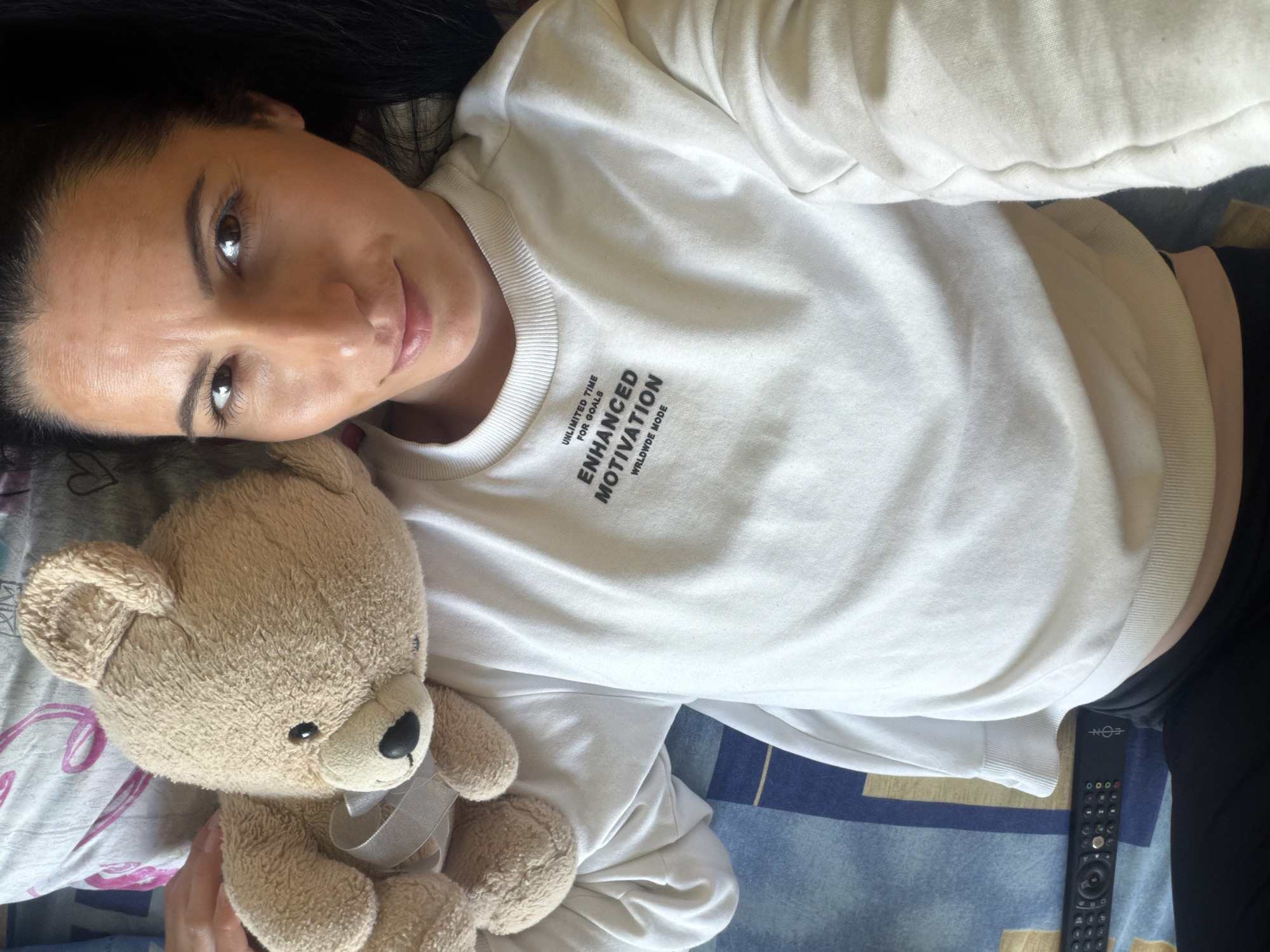 A woman lies on a bed with a blue, patterned bedspread. She is wearing a white sweatshirt that reads "Unlimited Time For Goals, Enhanced Motivation Worldwide Mode". She is holding a tan teddy bear with a silver ribbon. Next to her is a black remote control. The location is not discernible from the image.