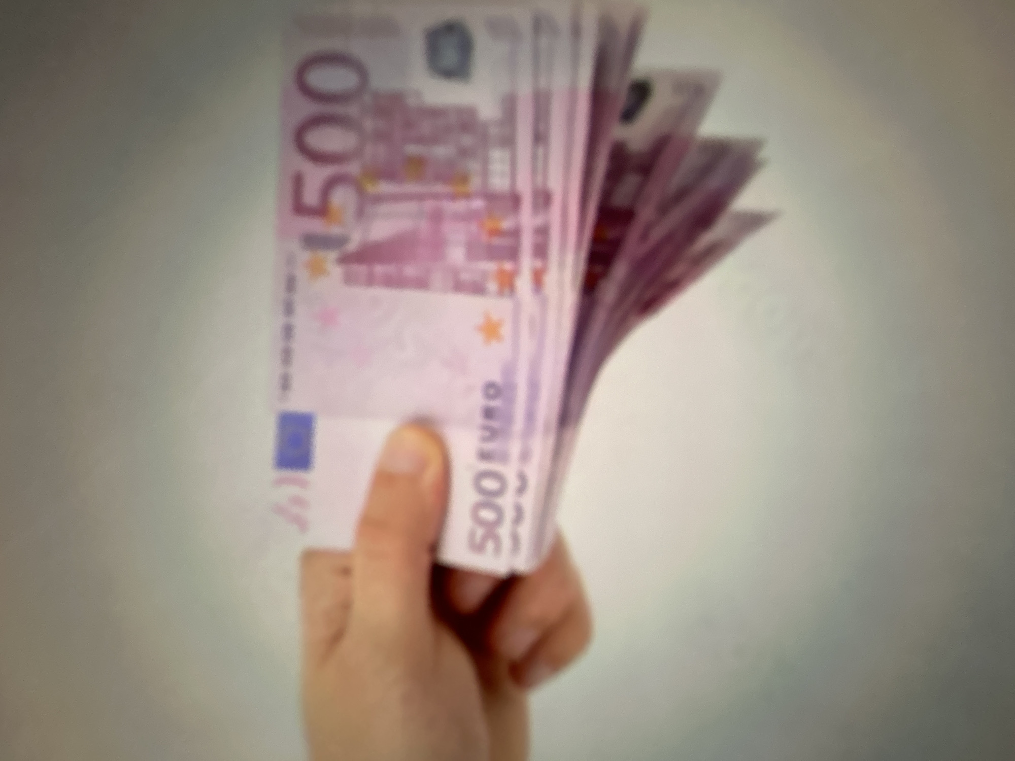 A hand holds a fan of 500 euro banknotes against a blurred background. The banknotes are predominantly pink and purple, with visible serial numbers and security features. The image is out of focus, making it difficult to discern finer details on the currency. The lighting appears to be artificial and diffused. The scene is indoors, with no identifiable external environmental cues. The overall mood is neutral, focusing on the presentation of the money. There is no indication of a specific time of day or weather conditions. The currency visible is clearly marked with "500 EURO." The provided location context of Gelendzhik, Russia, does not appear to be visually supported by any elements within the image itself.