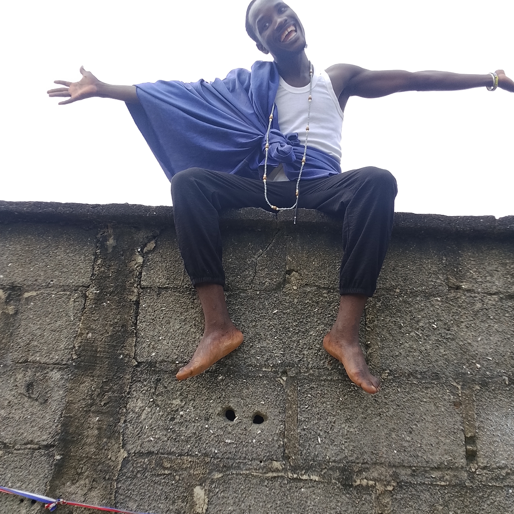A young man, appearing to be of African descent, is sitting on a textured concrete wall. He is wearing a white tank top and black pants. A blue garment is draped over his left shoulder and across his chest. He has a string of beads around his neck. His arms are outstretched to his sides. His feet are bare and dangling over the edge of the wall. The background is a bright, overcast sky. The wall appears to be part of an outdoor structure, possibly a fence or building, with visible lines and texture indicating it is constructed from concrete blocks. The location is identified as Ado, Nigeria.