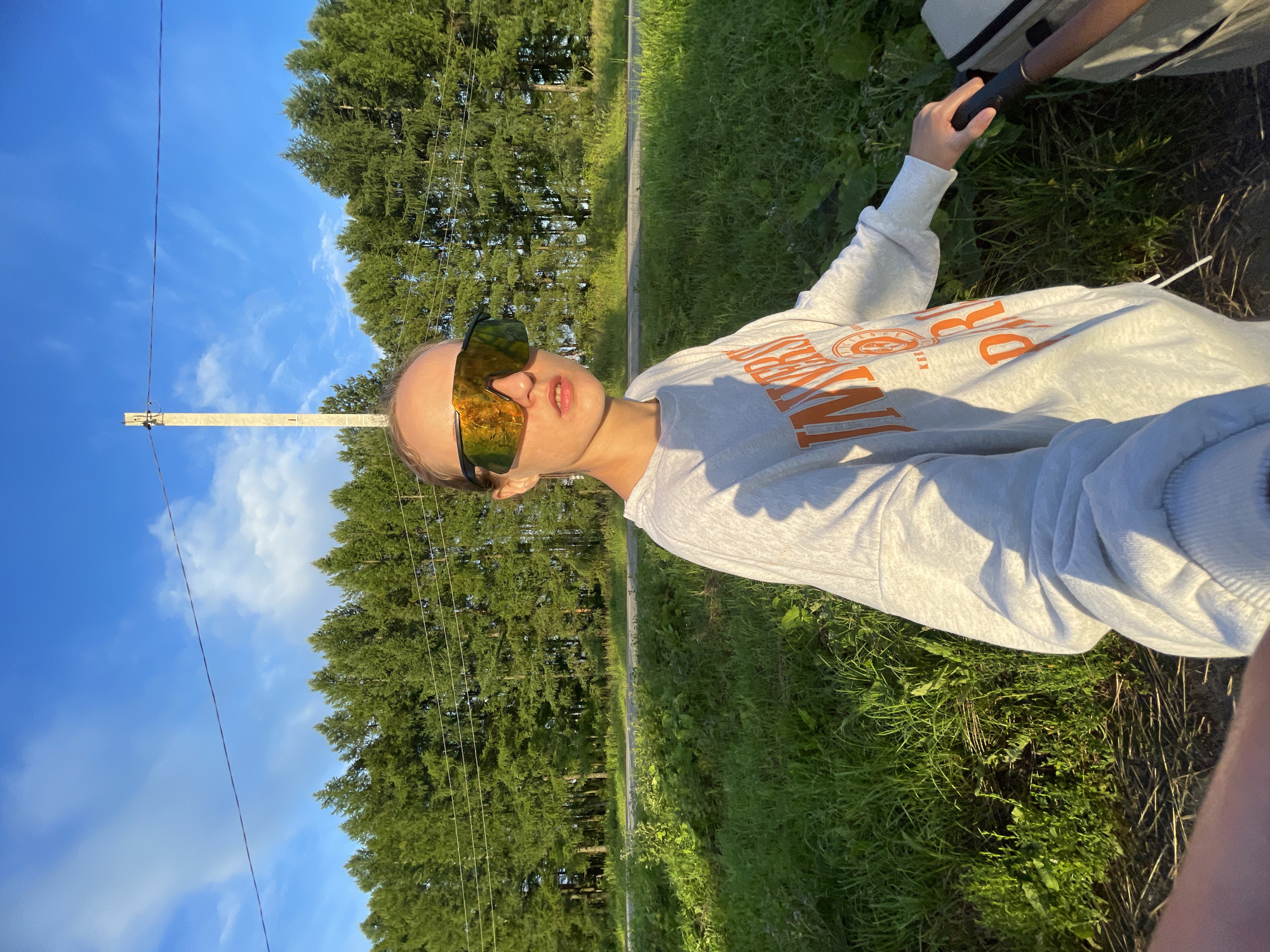 A young woman wearing oversized, reflective sunglasses and a light grey sweatshirt with orange lettering stands on a dirt path. The sweatshirt has the word "UNIVERSITY" emblazoned across the front in a distressed, vintage style. She is holding onto the handle of a stroller, with only the top portion of the stroller visible. The woman is looking directly at the camera. Behind her, a dense line of evergreen trees forms a backdrop, with a bright blue sky and scattered clouds above. A utility pole with wires runs through the scene. The lighting suggests it is either early morning or late afternoon, with the sun casting long shadows. The environment appears to be rural or semi-rural, possibly on the outskirts of a town. The woman is positioned slightly to the right of the frame, with the stroller handle extending from the right edge. The ground in the foreground is a mixture of dirt and dry grass. The overall impression is one of a casual outing, possibly a walk in the countryside.
