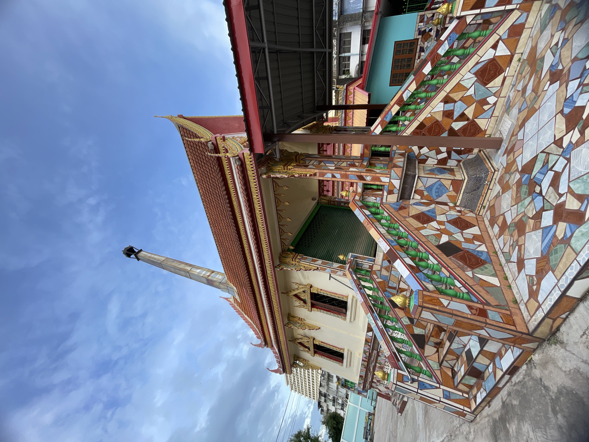 The image shows a cremation building in Pattaya City, Thailand, with a tall, weathered metal chimney extending from its roof. The building features a red-tiled roof with ornate golden trim and decorative elements. The exterior walls are cream-colored with yellow window frames and elaborately decorated window surrounds. A set of stairs leading up to the entrance is covered in a mosaic of colorful tiles in geometric patterns. The railings of the stairs are painted green and topped with golden finials. To the right of the cremation building, a modern structure with a corrugated metal roof is visible, also with a red trim. In the background, there are apartment buildings, one of which is tall and light-colored, and a blue wall. The sky is partly cloudy, with streaks of white clouds against a blue background. The ground in front of the building is paved with concrete and more tiled mosaics. There are no people visible in the image.