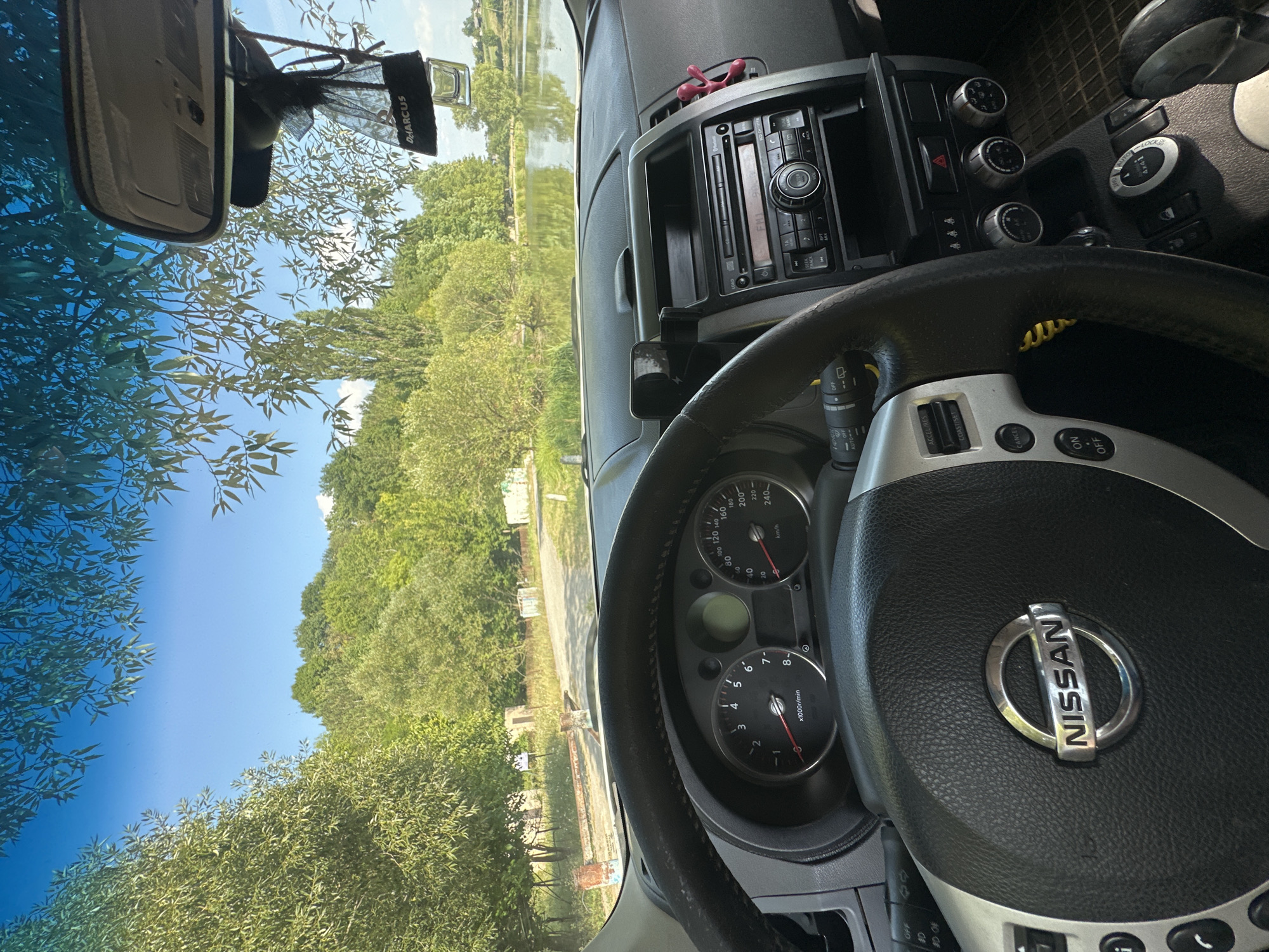 The image is a first-person perspective from inside a Nissan car, looking out through the windshield and side windows. The car is parked on an unpaved road, with a body of water visible in the background, likely a lake or pond. Lush green trees and foliage surround the water and extend up a hill. The sky is clear and blue. The interior of the car shows the steering wheel with the Nissan logo, the dashboard with a speedometer and tachometer, and the center console with a car stereo. A black air freshener hangs from the rearview mirror, and a phone mount is attached to the dashboard. In the distance, near the water's edge, there are some low structures, possibly old buildings or sheds. The overall impression is a peaceful, natural setting, likely in the countryside near Makiv, Ukraine, given the context provided.