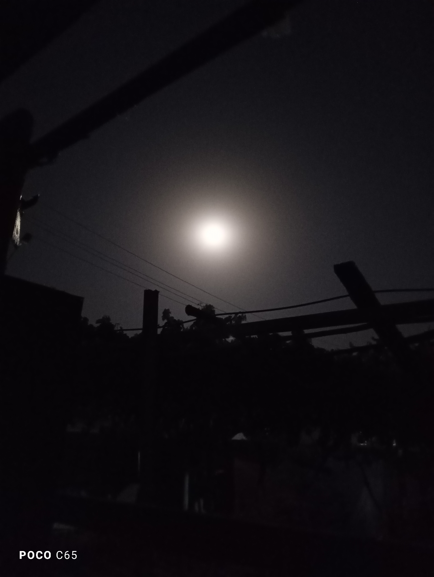 The image shows a nighttime scene dominated by a bright, full moon in a dark sky. Silhouetted against the moonlight are the indistinct shapes of trees, overhead wires, and some man-made structures, possibly part of a building or an outdoor shelter. The overall impression is one of a quiet, possibly rural or semi-urban environment at night. There is no indication of the city of Balkh, Tajikistan, or any specific geographical markers within the image. The foreground is very dark, obscuring details. The bottom left corner of the image displays the text "POCO C65".