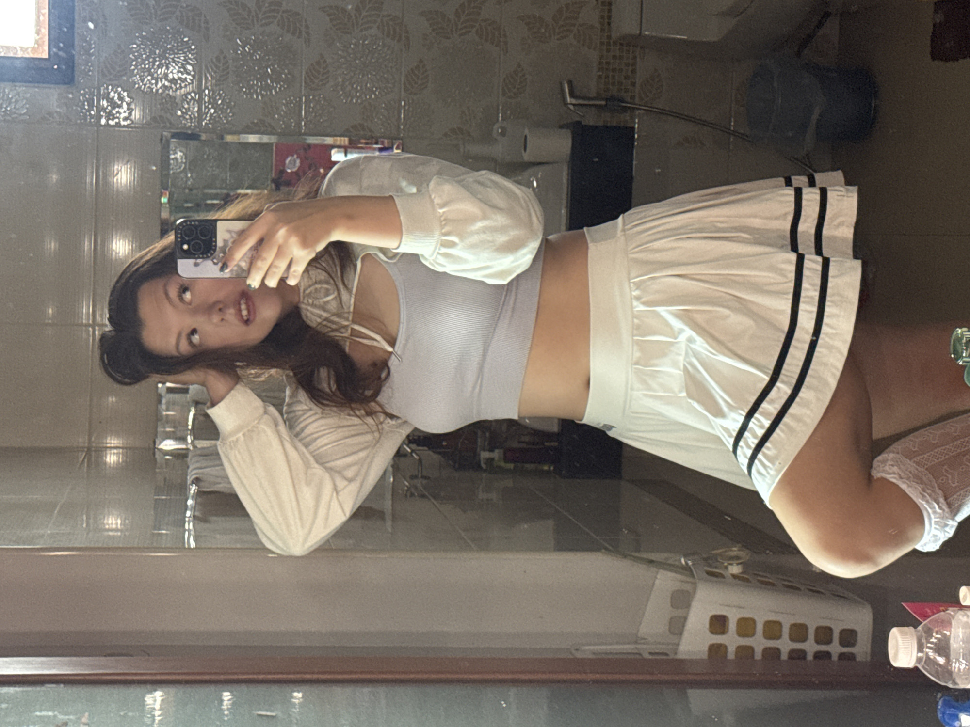 A young woman is taking a mirror selfie in what appears to be a bathroom. She is wearing a white long-sleeved crop top with a light blue ribbed halter top underneath and a white pleated skirt with black stripes. She has her hair up in a bun and is looking into the mirror with her phone held up. The bathroom has tiled walls with a floral pattern and a vanity with various bottles and a hamper. The location is identified as บ้านกาญจน์กนก 23, Thailand.