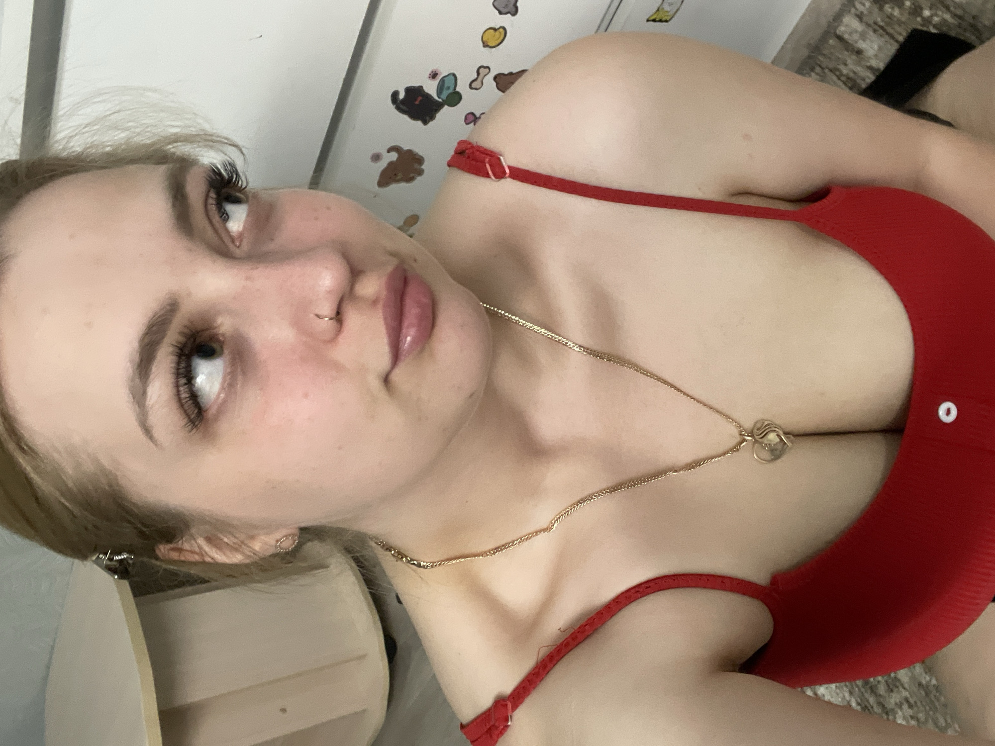 A close-up, angled selfie depicts a young woman with fair skin, light blonde hair, and prominent eyelashes. She has a nose piercing and is wearing lip gloss. She is wearing a red tank top and a gold necklace with a pendant. The background includes a white surface with cartoon-like stickers and a partial view of a light-colored piece of furniture. The location, Izhevsk, Russia, is not discernable from the image.