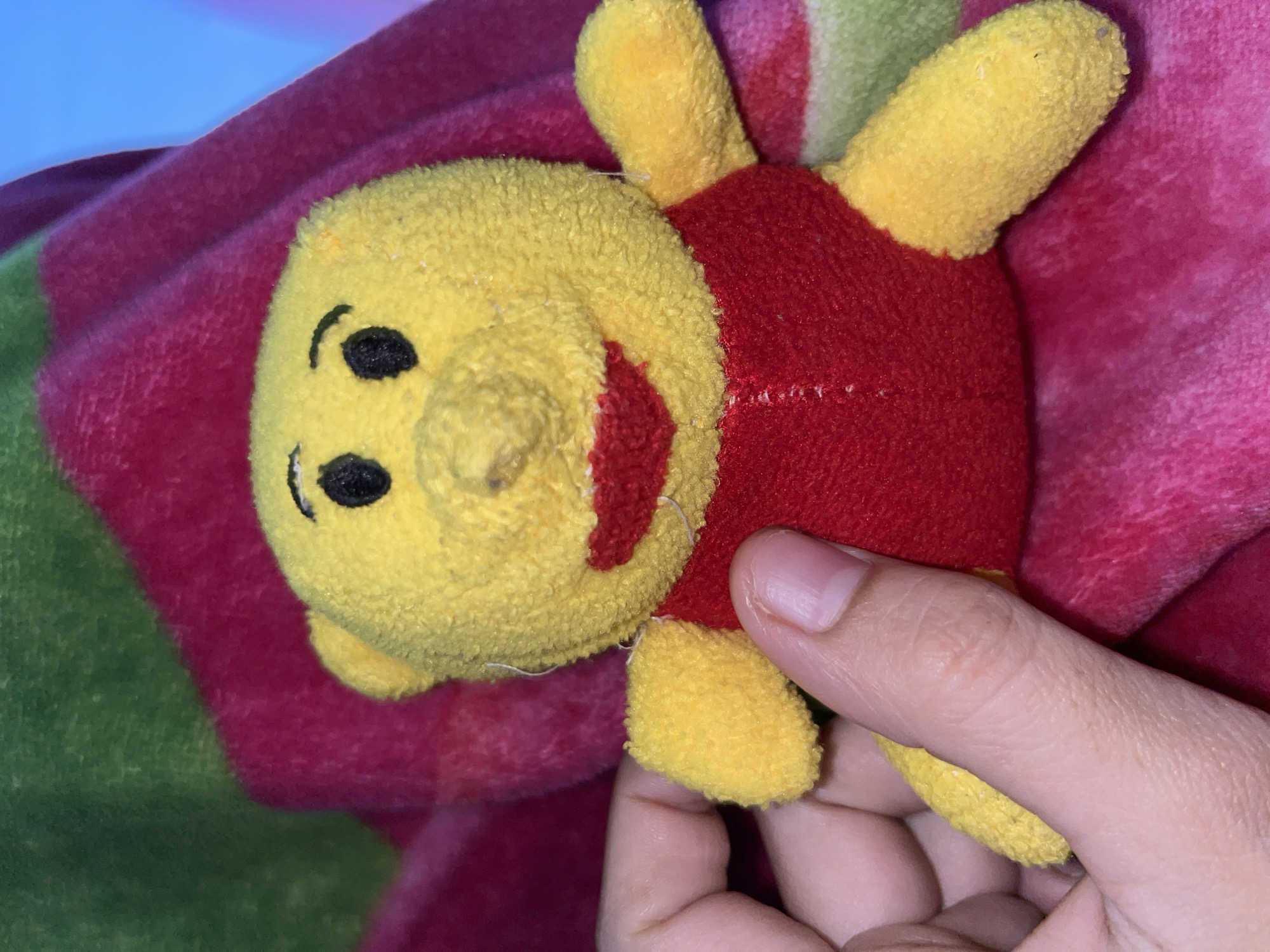 A close-up shot features a small, soft plush toy, strongly resembling Winnie the Pooh, held by a hand. The toy is predominantly yellow with a red body. It has embroidered black eyes, a small yellow nose, and a red, smiling embroidered mouth. Its four short, stubby limbs are also yellow. The plush material appears slightly worn and fuzzy. A hand with light brown skin is visible on the right side of the frame, gently holding the toy's lower body and one of its limbs.
The background consists of a soft, textured fabric, likely a blanket or throw, which displays broad stripes or sections of deep red, bright pink, and muted green. The fabric is slightly rumpled. The lighting is soft and even, suggesting an indoor setting without direct sunlight. There are no specific cues for time of day or weather, nor is any text visible in the image. The scene conveys a simple, domestic moment in Lhokseumawe, Indonesia.