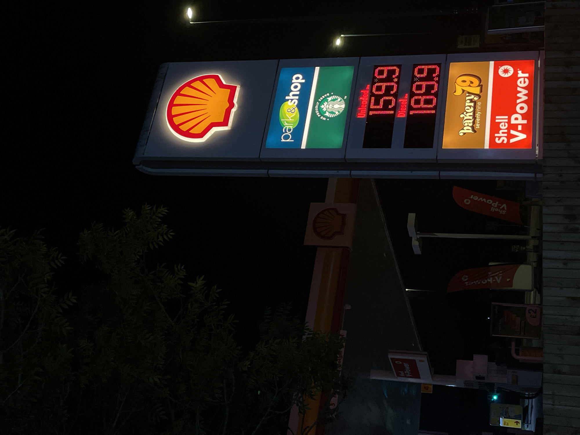 The image shows a Shell gas station at night. The sign displays the Shell logo, along with "park&shop," and "We Proudly Serve" with a Starbucks logo. The price for unleaded gasoline is shown as 159.9 and diesel fuel is 189.9. There is a "bakery 79 seventy nine" sign and "Shell V-Power" is also displayed on the sign. The surrounding environment includes a tree and other gas station signage. Maidstone, United Kingdom is not specified in the image.