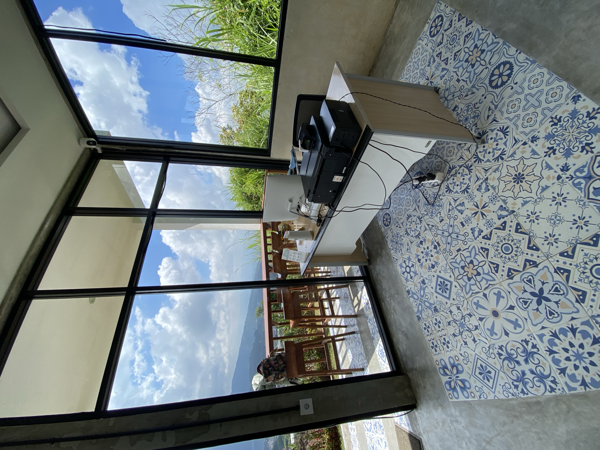 A security camera is mounted on the corner of a ceiling in a modern room with large glass windows offering a view of a lush green hillside and a blue sky with white clouds. Inside the room, a white desk is set up with a computer monitor, a printer, and a scanner. A power strip with several plugs is attached to the desk leg. A patterned rug covers a portion of the floor. In the background, beyond the glass windows, there is an outdoor seating area with wooden bar stools and a railing, overlooking a mountainous landscape.