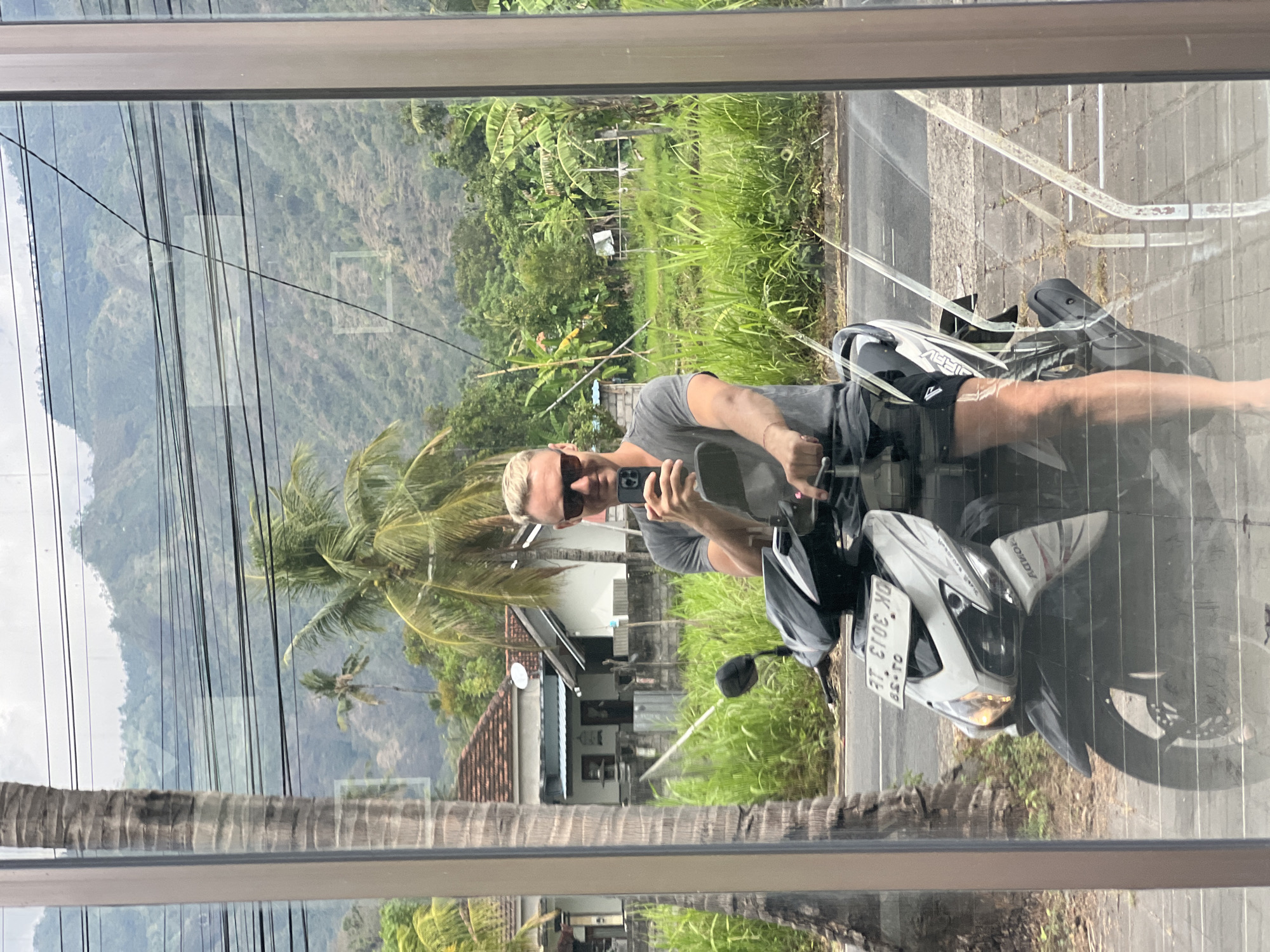 A man with blonde hair and sunglasses is seated on a white and black scooter, holding a phone in his right hand to take a selfie. He is wearing a grey t-shirt and black shorts. The scooter has a license plate that reads "AT 6106 XD". Behind him, a palm tree trunk is visible to the left, and further back, houses with red tile roofs and lush green vegetation can be seen. In the background, rolling hills covered in greenery and a cloudy sky are present. The scene appears to be in a rural or semi-rural area of Abang, Indonesia, possibly on a street with a reflective surface like a window or a car.
