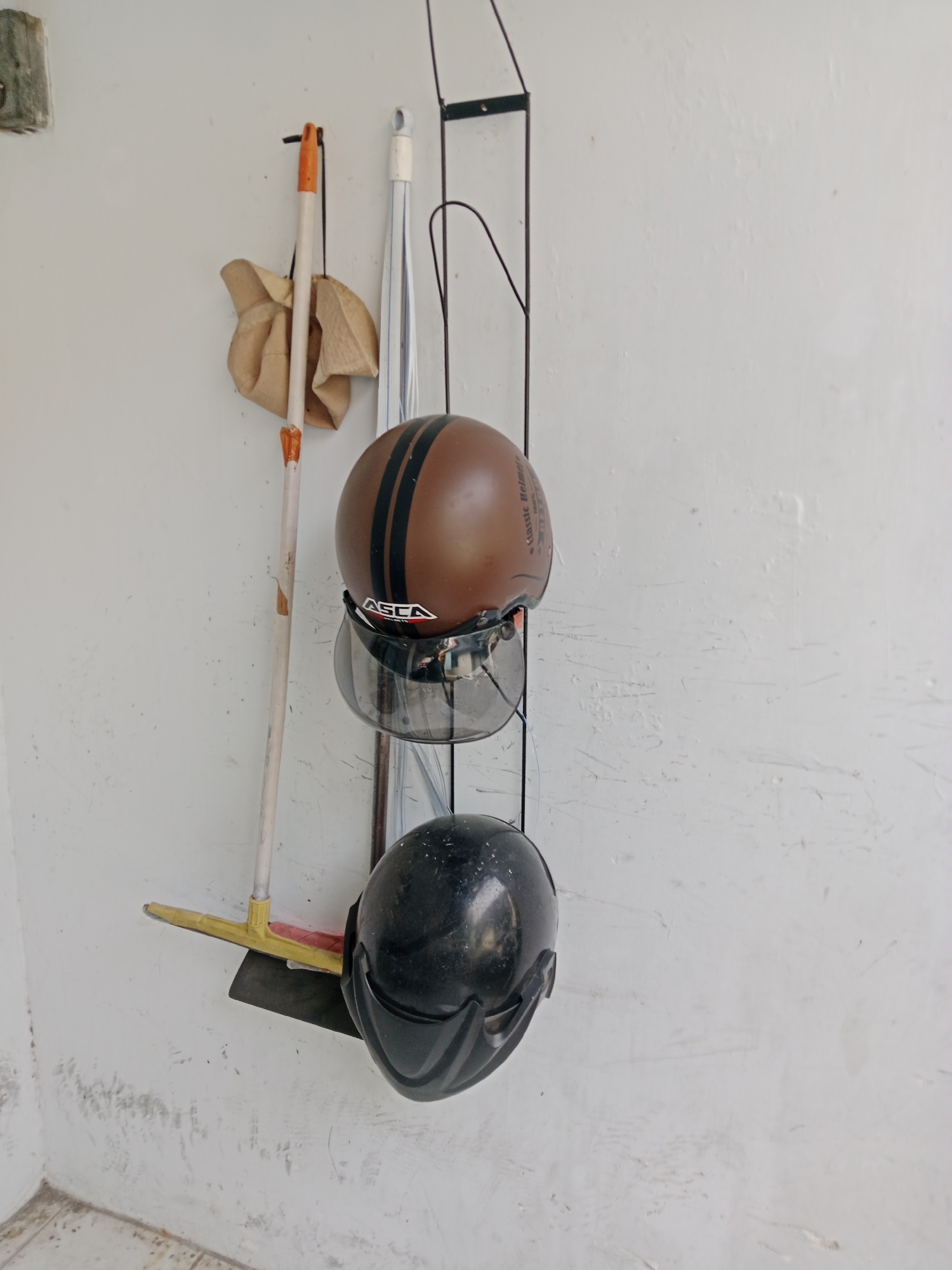 Two motorcycle helmets, one brown with black stripes and a clear visor, and one black with a tinted visor, are hanging on a black metal rack mounted on a textured white wall. Below the helmets, a broom with a yellow handle and a black dustpan are leaning against the wall. To the left of the broom, a rag or cloth is draped over another object. The scene appears to be an outdoor storage area or garage, possibly in Kediri City, Indonesia, given the context. The wall shows signs of wear and tear, with scuff marks and uneven texture. The floor visible at the bottom of the image is tiled.