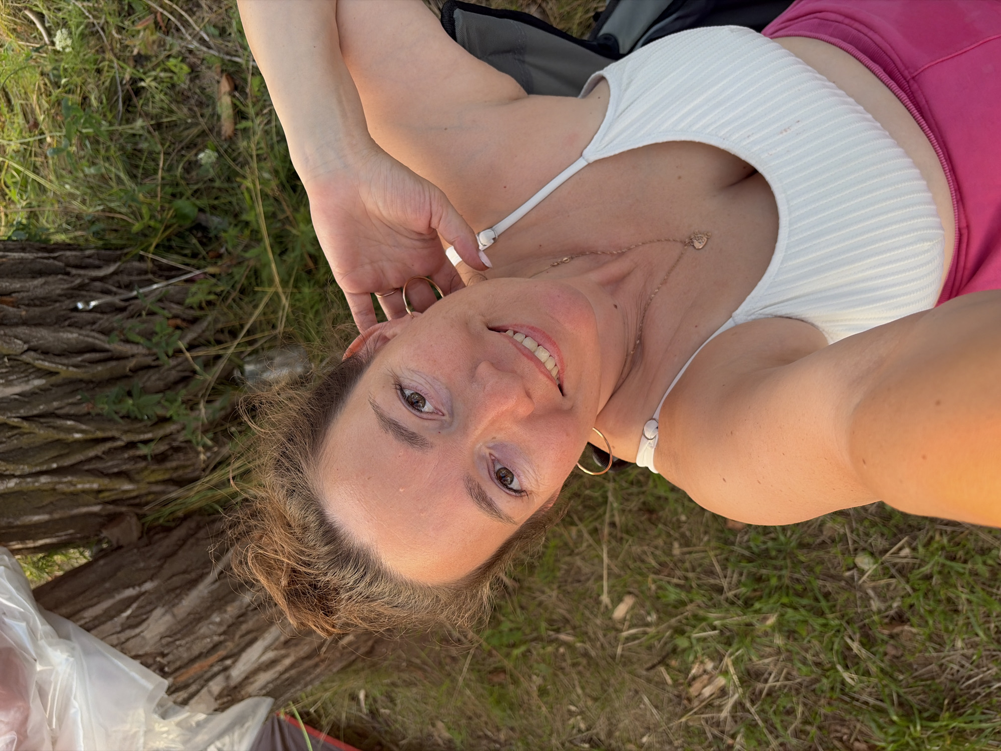 The media file shows a woman lying on her back in a grassy outdoor setting. She is wearing a white ribbed tank top and pink shorts. Her head is tilted back, and she is smiling at the camera, with her eyes open and looking forward. Her right hand is raised and touches her hair. She is wearing gold hoop earrings and a delicate necklace with a heart-shaped pendant. The background consists of grass, tree bark, and some foliage. A white plastic bag and part of a tent are visible in the lower left corner. The lighting suggests it is daytime, possibly late afternoon due to the warm tones. The overall mood appears relaxed and happy. The location is Ol'khovskoye, Russia. There is no visible text in the image.