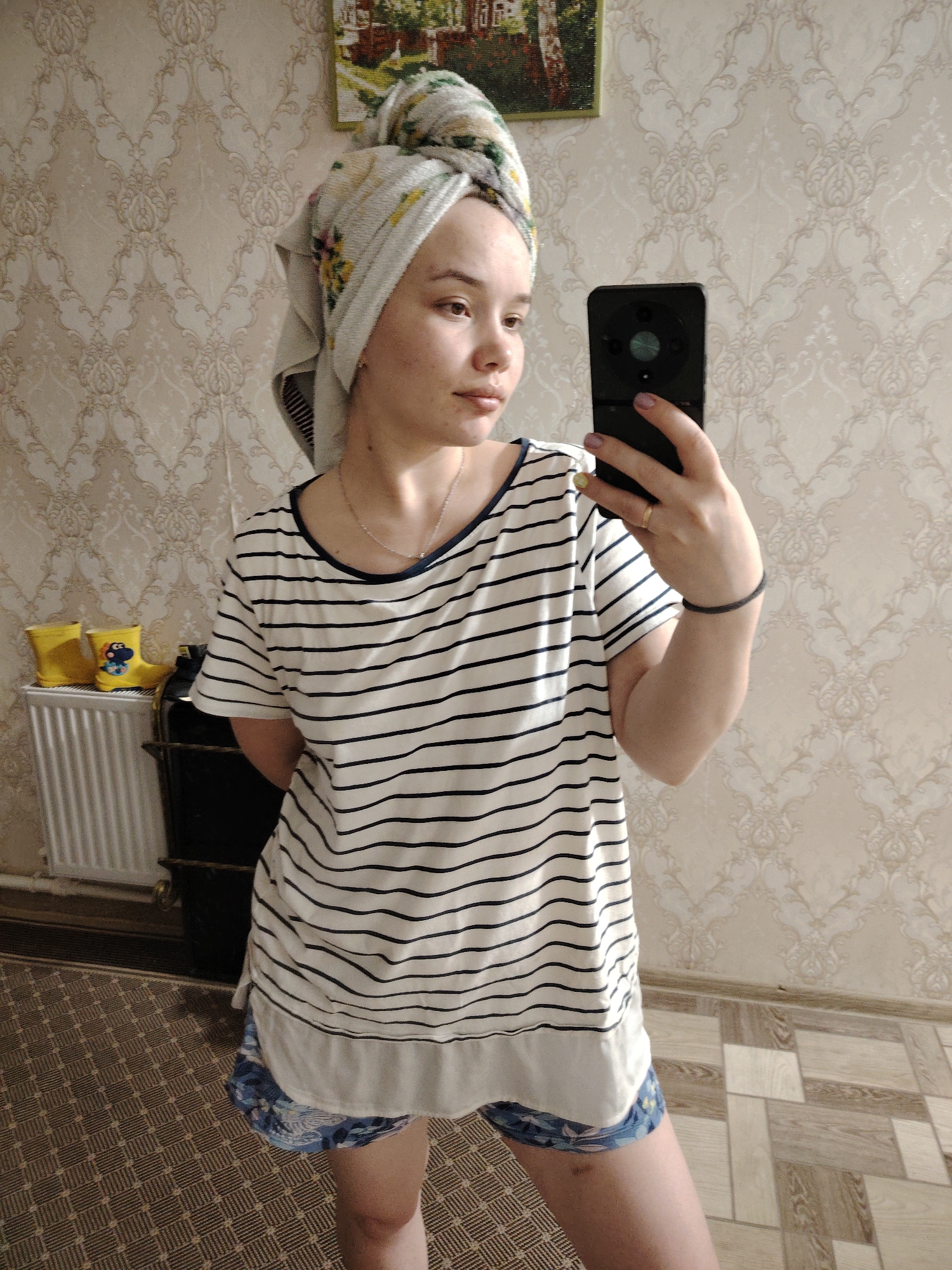 A young woman stands indoors, holding a black smartphone in her right hand, taking a selfie in front of a wall with patterned beige wallpaper. She has a towel wrapped around her head and is wearing a white and navy striped t-shirt with a white panel at the bottom, and blue patterned shorts. She wears a ring on her left hand and a black band on her right wrist.
To the left of the woman is a white radiator with yellow rubber boots placed on top. A black storage unit is beside the radiator. The floor is covered in patterned linoleum. A framed picture hangs on the wall above the woman.
It is impossible to establish the location in the city of Noviy, Russia from this image.