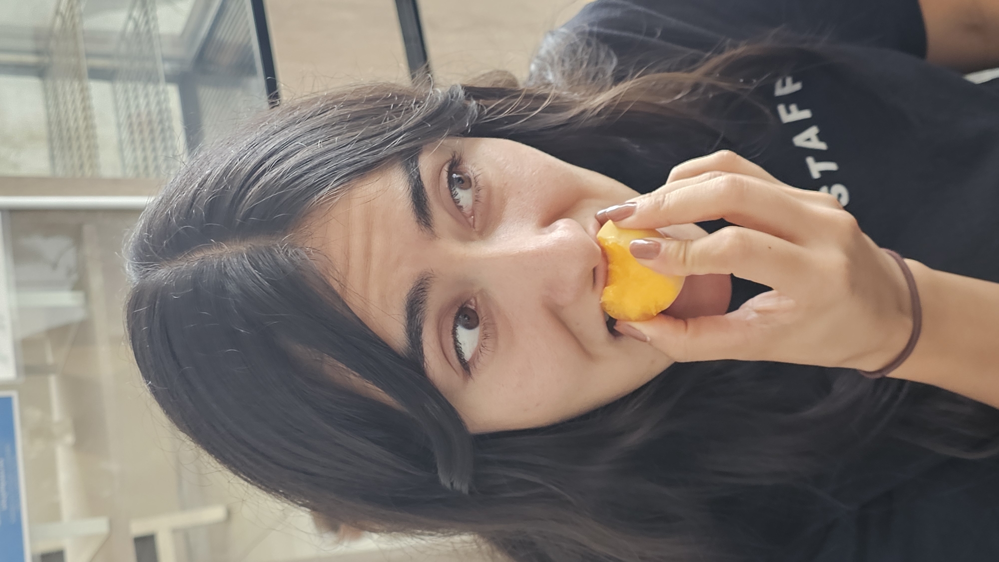 A young woman with long, dark hair is depicted in a close-up, chest-up shot, looking upwards and slightly to her right. She holds a partially eaten, light orange fruit, resembling a peach or nectarine, to her mouth with her right hand, suggesting she is actively eating it. Her brown eyes are open, and her expression is neutral, possibly looking at the camera or an unseen subject above it.
She is dressed in a dark-colored (likely black) t-shirt, with the white capital letters "STAFF" partially visible on the upper right side of her chest. A dark brown elastic hair tie adorns her right wrist, and her fingernails are painted a light brown or nude color.
The background is softly out of focus, revealing an indoor or semi-outdoor setting. Large windows or glass panels with a subtle grid-like pattern are visible, alongside some lighter, indistinct structures in the distance, possibly architectural elements or furniture. The bright lighting suggests it is daytime. The scene is situated in Puebla, Mexico.