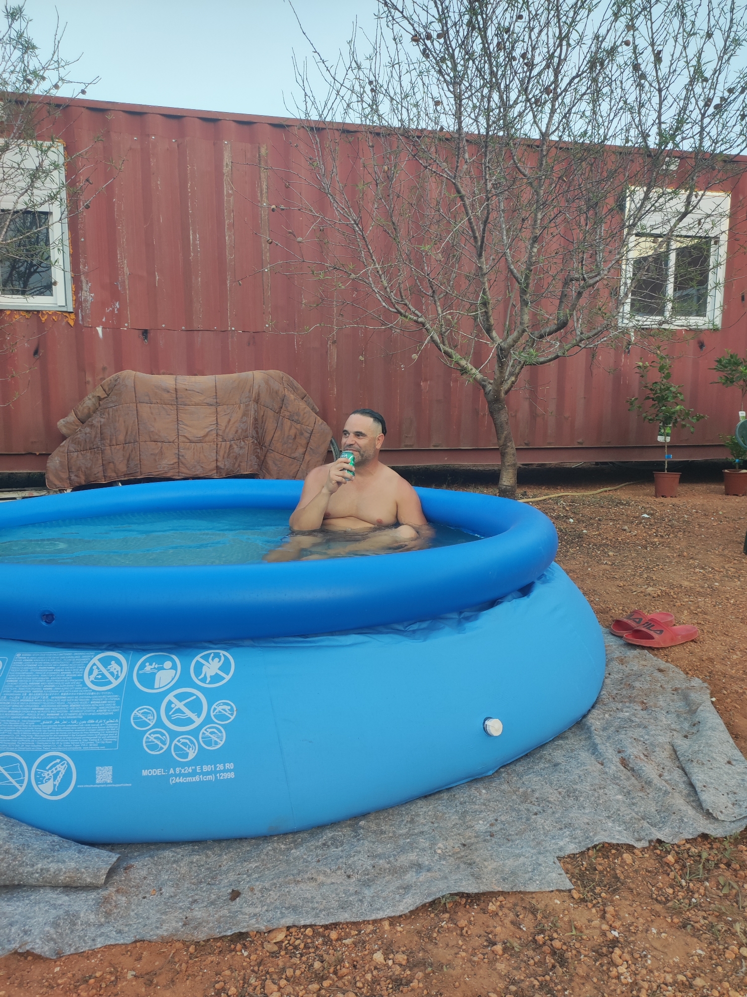 A man is sitting in a blue inflatable pool filled with water, holding a can in his right hand and drinking from it. He is shirtless and appears to be relaxing. The pool is situated on a patch of dirt and gravel, with a gray tarp underneath it. In the background, there is a large, rusted red shipping container that has been converted into a structure with windows. A bare tree stands behind the pool, and a few small potted plants are visible to the right. Red flip-flops are on the ground near the pool. The scene is set outdoors, likely in a rural or semi-rural area, consistent with a property in Alcàsser, Spain.